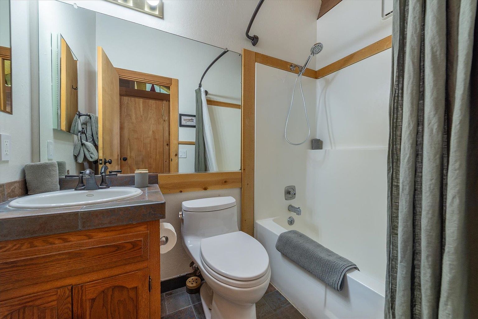 This compact bathroom features a vanity with a dark stone countertop and a wood cabinet, paired with a white toilet and a shower-tub combination. A large mirror spans the wall above the vanity, reflecting the wooden door and the shower area, which is enclosed by a curtain. The space is accented with wood trim and neutral-toned walls, creating a functional and rustic aesthetic.