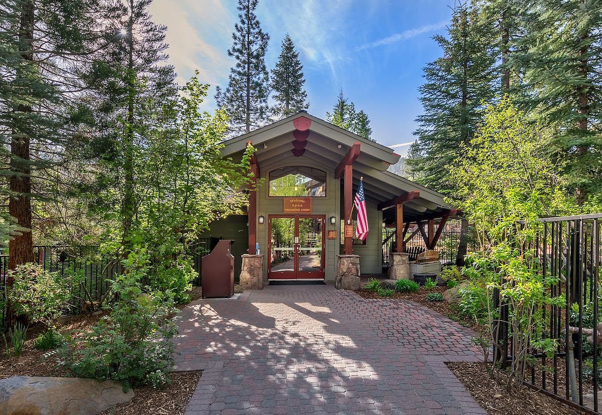 This image captures the inviting entrance of a recreation center, featuring a covered porch with timber framing and stone-clad pillars. A brick-paved walkway leads directly to the glass-paneled double doors, which are flanked by lush greenery and tall pine trees. The scene is bathed in bright, natural daylight, creating a welcoming and serene atmosphere for a community facility.