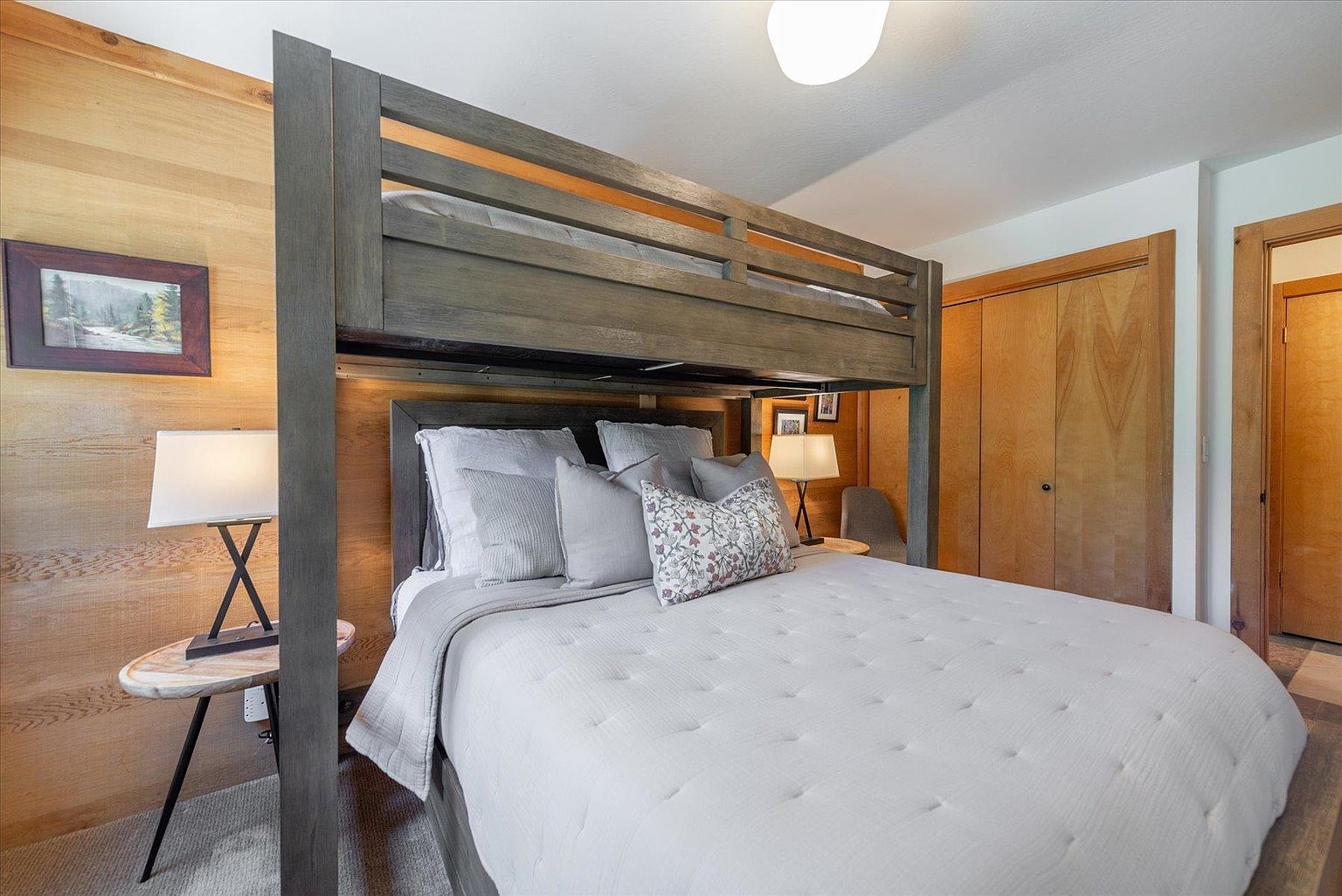 This cozy bedroom features a rustic aesthetic with light wood-paneled walls and a sturdy, dark-toned wooden bunk bed frame. The lower bed is dressed in a neutral, quilted bedding set with a variety of decorative pillows, while two small side tables with lamps provide warm accent lighting. The room includes a wooden closet door and a framed landscape print, creating a comfortable and inviting atmosphere.