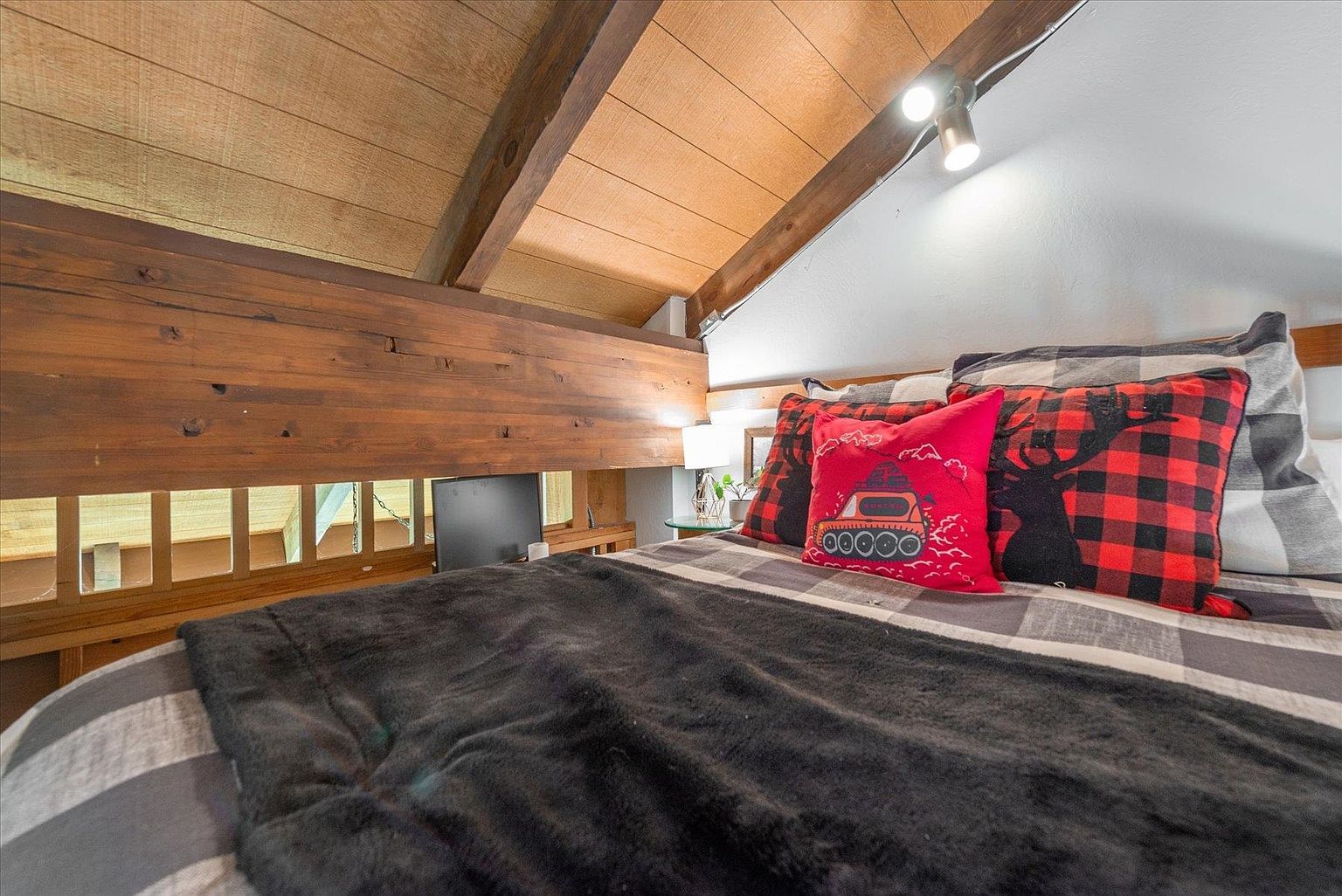 This cozy loft bedroom features a rustic aesthetic with exposed wooden beams and wood-paneled walls. The bed is dressed in a plaid comforter and accented with a festive red pillow, creating a warm, cabin-like atmosphere. A small spotlight mounted on the ceiling provides focused lighting, while a wooden railing overlooks the space below.