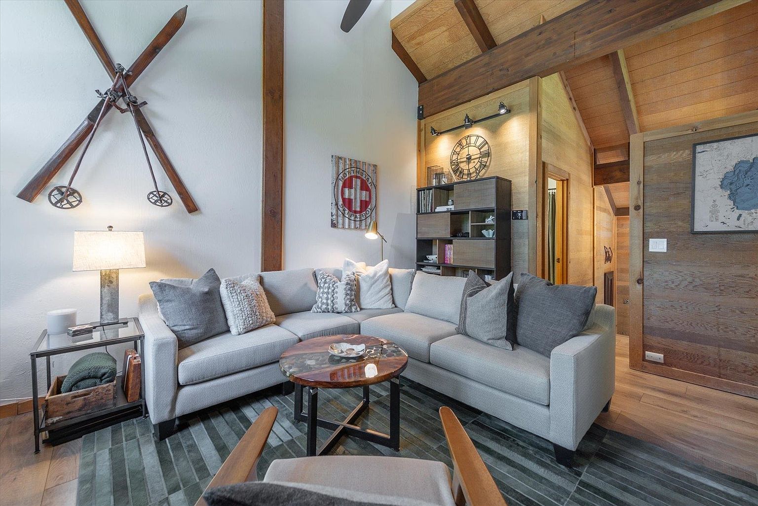 This cozy, mountain-inspired living room features a large, light-toned sectional sofa arranged around a circular wood and metal coffee table. The space is accented with rustic decor, including vintage-style skis mounted on the wall and a Northstar Ski Patrol sign, all set against a backdrop of exposed wood beams and high, vaulted ceilings. A dark wood shelving unit and a large wall clock add character to the room, which is illuminated by warm, ambient lighting.