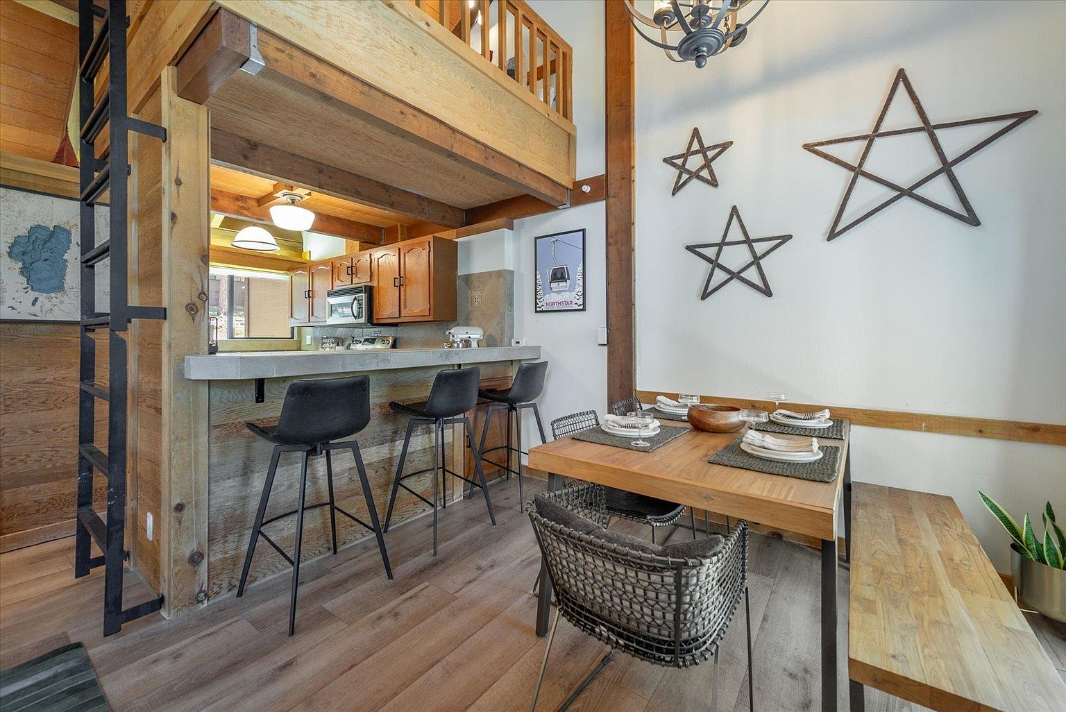 This open-concept dining and kitchen area features a rustic, mountain-inspired aesthetic with exposed wood beams and light wood flooring. A dining table with a bench and chairs sits adjacent to a breakfast bar with three stools, creating a functional space for meals. The room is decorated with metal star wall art and a framed ski-themed print, while a lofted area above the kitchen adds architectural interest.