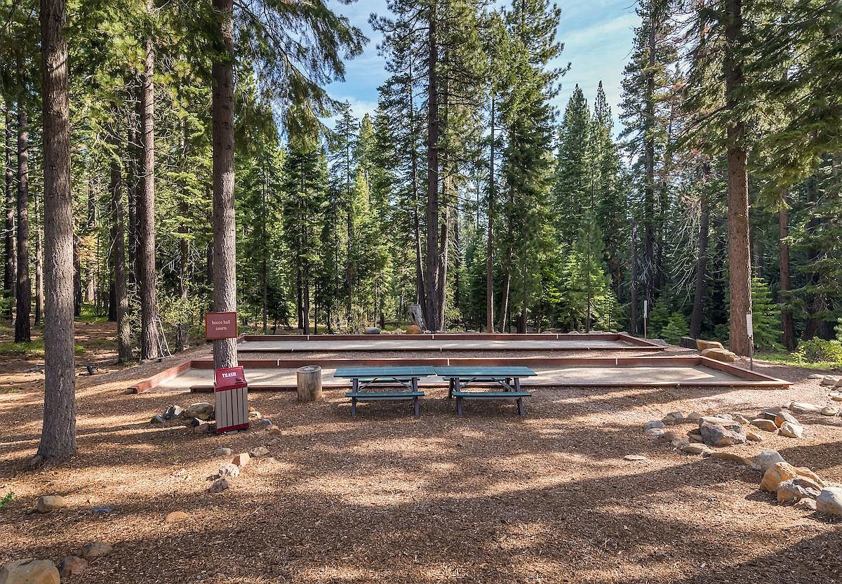 This outdoor recreational area features two side-by-side bocce ball courts nestled within a serene, wooded setting. A picnic table sits in the foreground on a bed of pine needles, providing a casual seating area for residents or guests. The scene is framed by tall, mature pine trees, creating a peaceful and shaded environment for community leisure.
