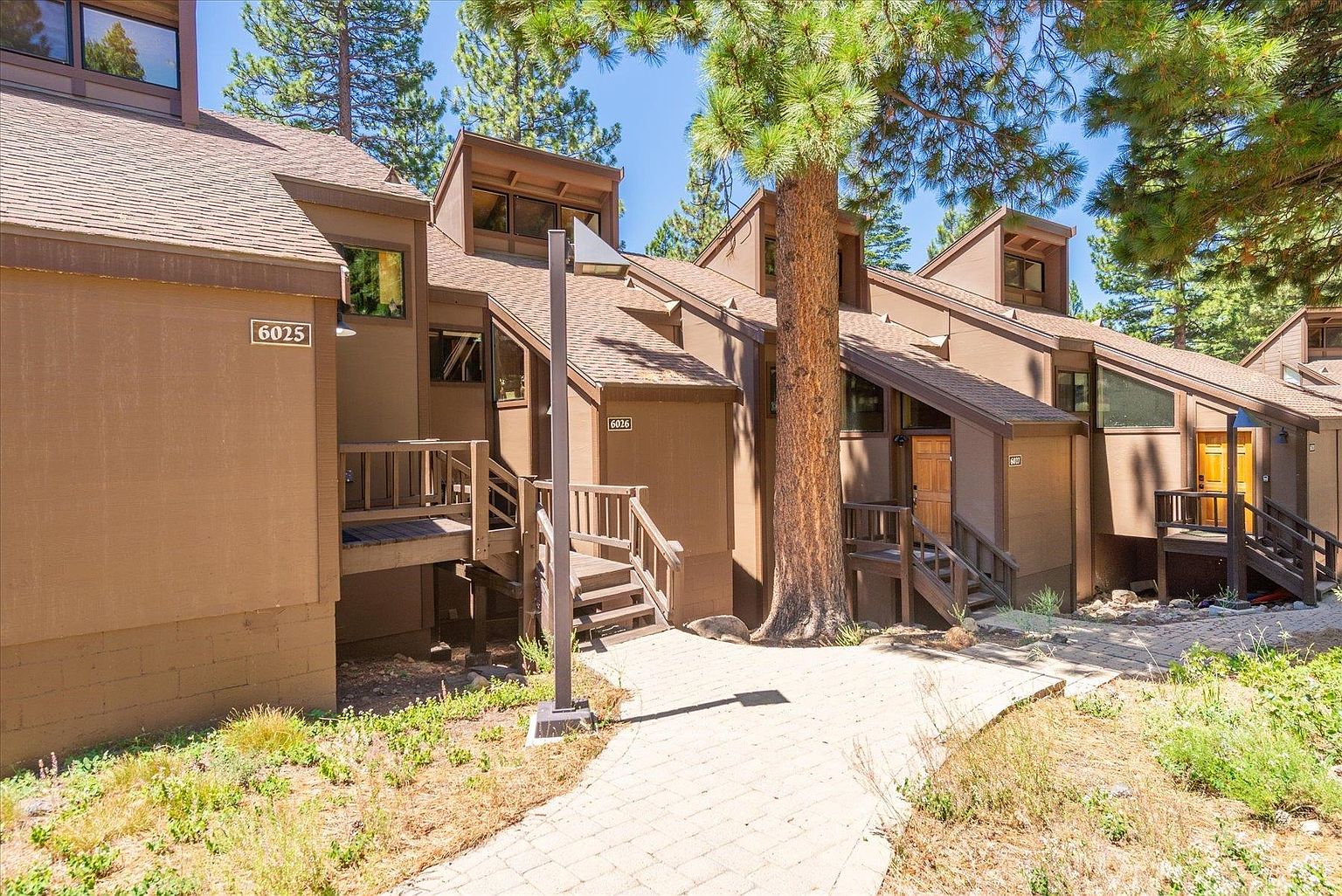 This exterior view captures a multi-unit residential complex featuring brown-toned siding and a series of gabled rooflines. A stone-paved walkway leads toward the wooden staircases and entryways of units marked 6025, 6026, and 6027, all nestled among mature pine trees. The perspective is at eye level, emphasizing the rustic, mountain-style architecture and the natural, wooded setting.