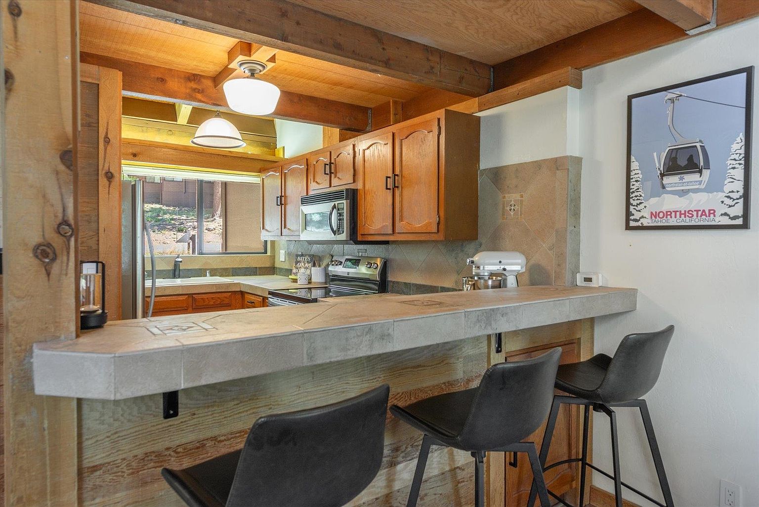 This kitchen features a rustic aesthetic with warm wood cabinetry, exposed ceiling beams, and a tiled breakfast bar that separates the cooking area from the living space. Three modern black bar stools are positioned at the counter, while a Northstar Tahoe poster adds a local touch to the wall. The space is illuminated by pendant lighting and natural light streaming through a window above the sink, creating a cozy, mountain-lodge atmosphere.