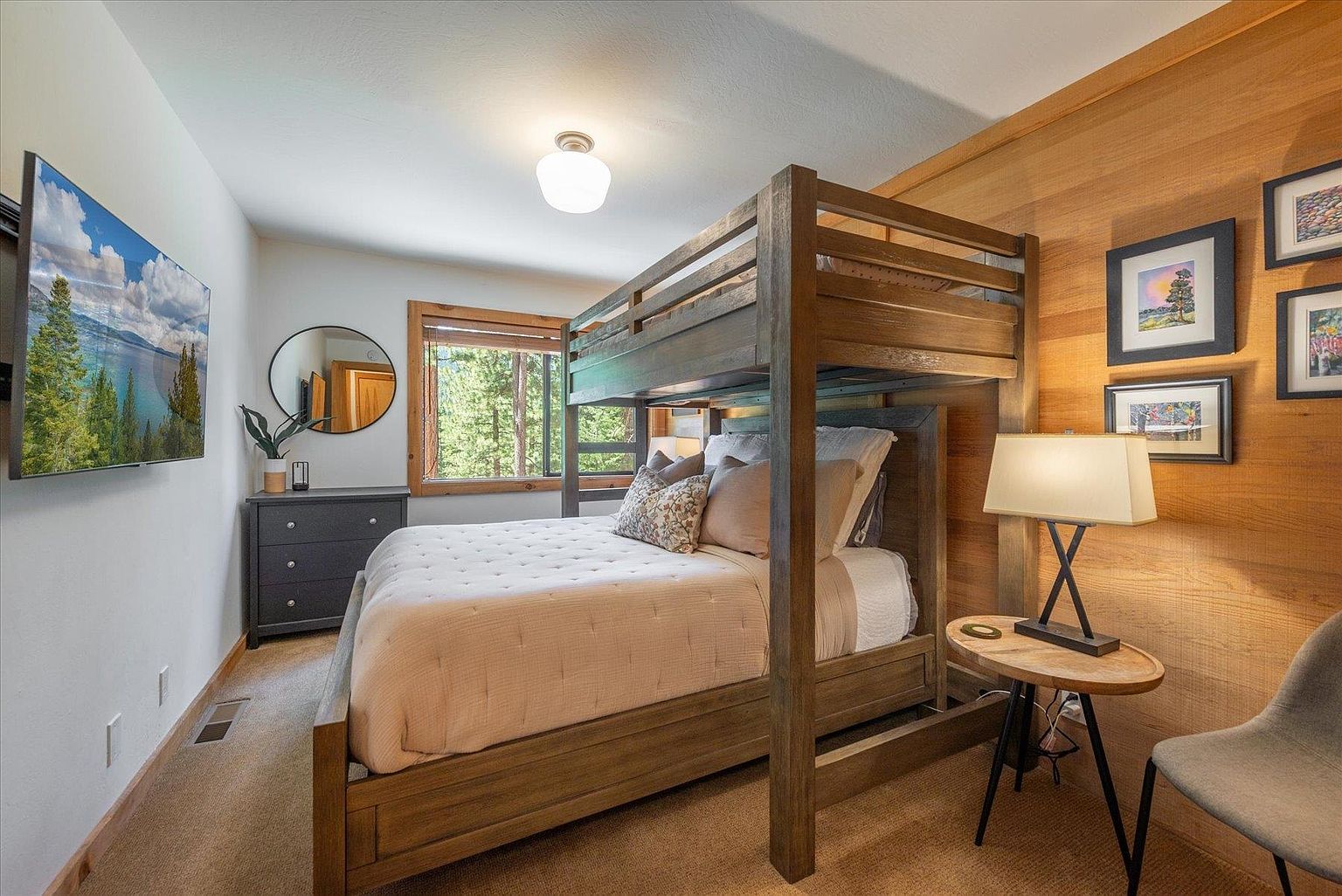 This cozy bedroom features a rustic wooden bunk bed set against a warm wood-paneled accent wall adorned with framed artwork. A dark dresser sits beneath a circular mirror, while a bedside table with a lamp provides a functional touch next to the bed. The room is completed by a wall-mounted television and a neutral color palette, creating a comfortable and inviting atmosphere.