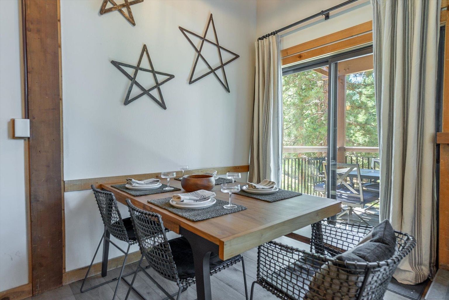 This cozy dining area features a rustic wooden table set for four, surrounded by modern wire-frame chairs. The space is accented by decorative metal star wall art and is bathed in natural light from the adjacent sliding glass doors that lead to an outdoor deck. The warm wood tones and clean lines create an inviting, mountain-inspired atmosphere.