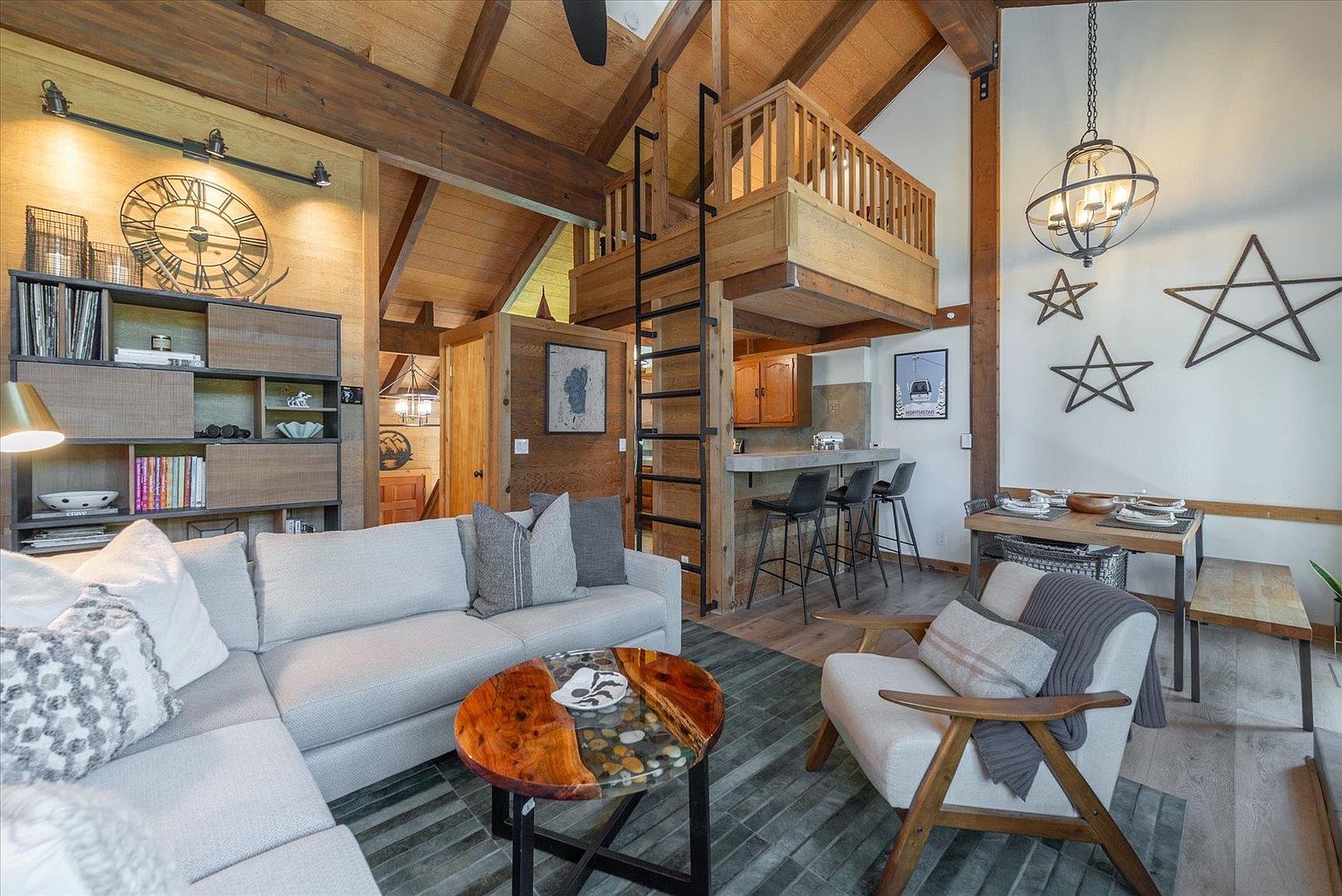 This cozy, rustic-inspired living room features a vaulted ceiling with exposed wooden beams and a loft area accessible by a black metal ladder. The space is furnished with a large, light-colored sectional sofa, a round wood-and-stone coffee table, and a comfortable armchair, all set against a backdrop of warm wood paneling and white walls decorated with star-shaped wall art. A dining area with a wooden table and bench is positioned to the right, while a small kitchen bar with stools sits beneath the loft, creating an open-concept layout perfect for mountain-style living.