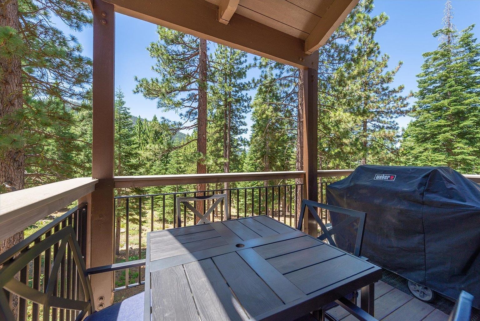This elevated deck offers a serene outdoor living space surrounded by a dense forest of tall pine trees. The area is furnished with a dark-toned dining table and chairs, complemented by a covered grill positioned to the side. The perspective captures the inviting atmosphere of a mountain retreat, emphasizing the connection between the private deck and the natural landscape.