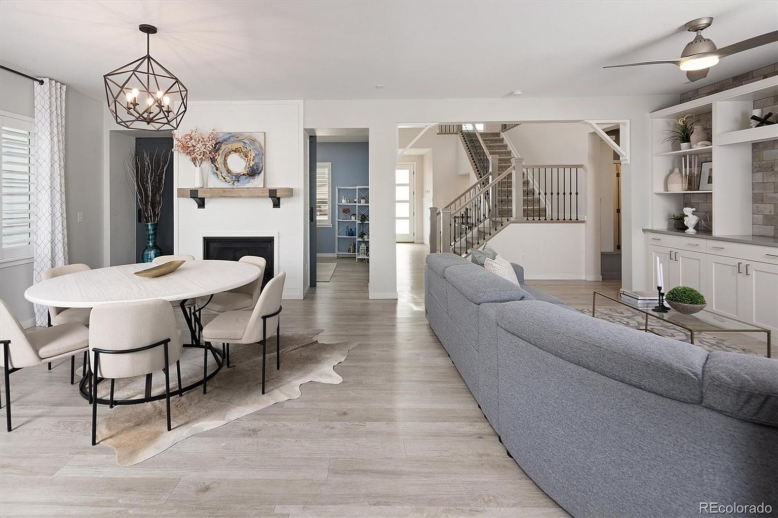 This open-concept living and dining area features a modern, airy aesthetic with light wood-look flooring and neutral tones. The space includes a cozy dining nook with a geometric chandelier and a fireplace, transitioning seamlessly into a spacious living room anchored by a large grey sectional sofa and built-in shelving. The perspective captures the flow of the home, highlighting the staircase in the background and the bright, inviting atmosphere.