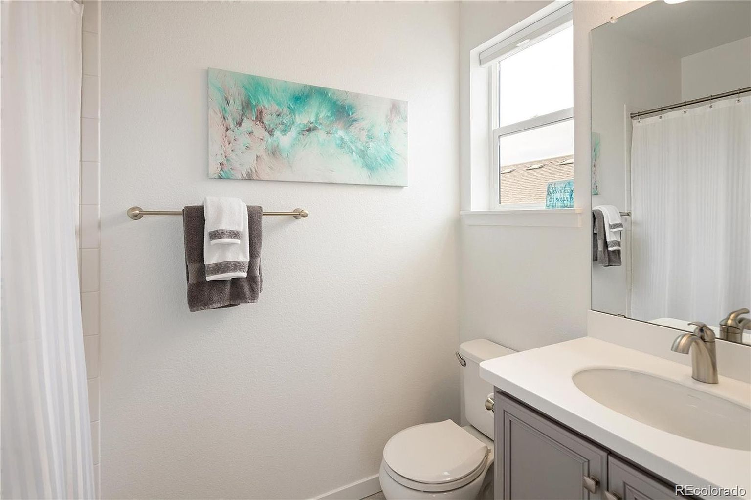 This clean and bright guest bathroom features a neutral color palette with white walls and a grey vanity cabinet. The space includes a standard toilet, a single-sink vanity with a large mirror, and a shower-tub combination with a white curtain. A piece of abstract wall art adds a touch of color, creating a welcoming and well-maintained atmosphere.