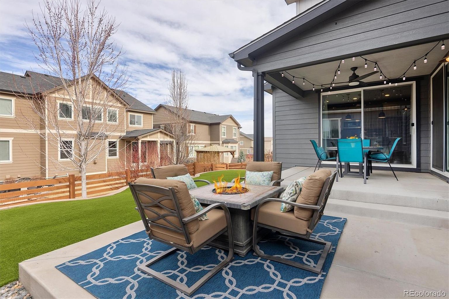 This inviting outdoor living space features a concrete patio furnished with a cozy fire pit set surrounded by four cushioned chairs on a patterned blue area rug. A covered patio area adjacent to the house includes a dining set and overhead string lights, creating a perfect transition to the well-manicured backyard. The perspective captures a comfortable, modern suburban lifestyle with a clear view of the surrounding neighborhood homes.