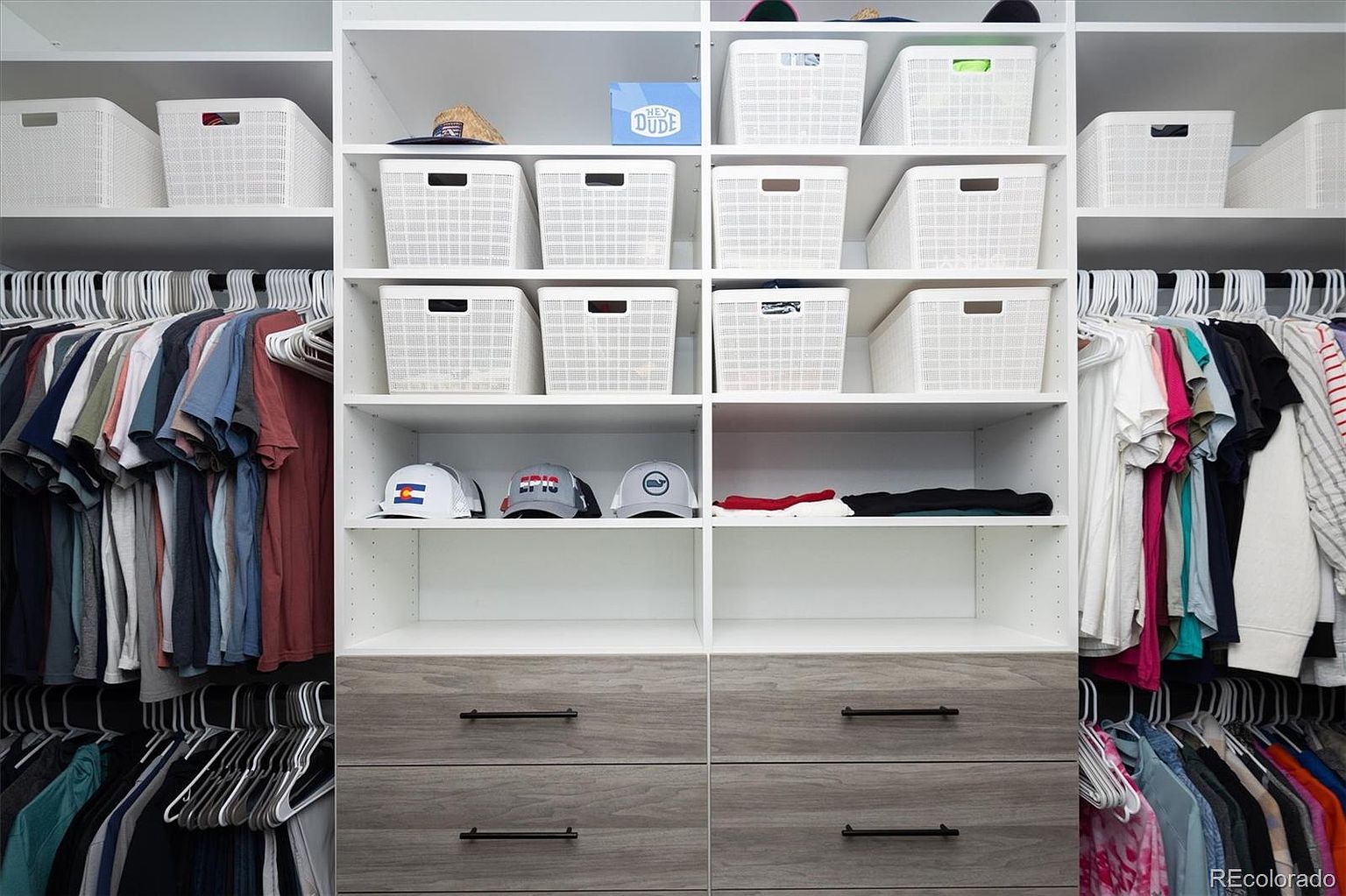 This organized walk-in closet features a clean, modern design with white shelving units and grey wood-grain drawers. The space is efficiently arranged with hanging rods for clothing on both sides and multiple white storage bins on the upper shelves, creating a tidy and functional aesthetic. The perspective is a straight-on, eye-level shot that highlights the ample storage capacity and neat organization of the closet.