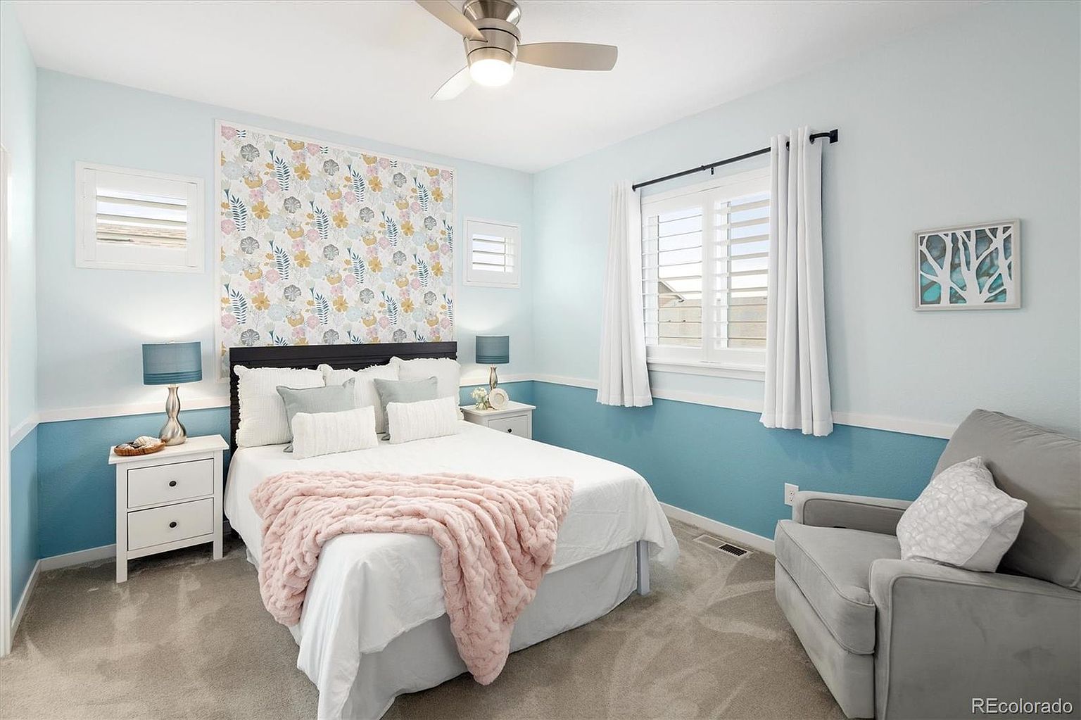 This bright and inviting bedroom features a queen-sized bed adorned with a soft pink throw blanket and a decorative floral wallpaper accent wall. The room is painted in a calming light blue with white wainscoting, complemented by a comfortable gray armchair and white bedside tables. The perspective is a straight-on shot capturing the cozy, well-lit atmosphere of the space.