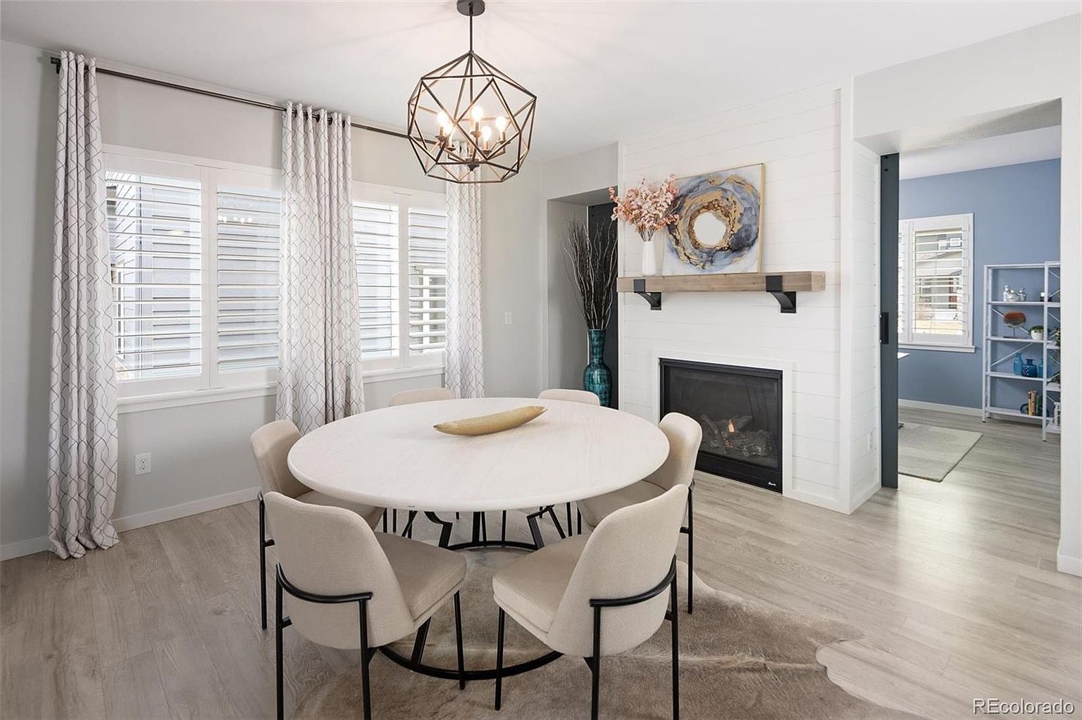 This bright and inviting dining room features a modern round table with upholstered chairs set atop a textured rug. A striking geometric chandelier hangs above the table, while a white shiplap fireplace with a rustic wood mantel serves as a focal point. The space is framed by elegant floor-to-ceiling curtains and offers a glimpse into an adjacent office, creating a cohesive and stylish open-concept feel.