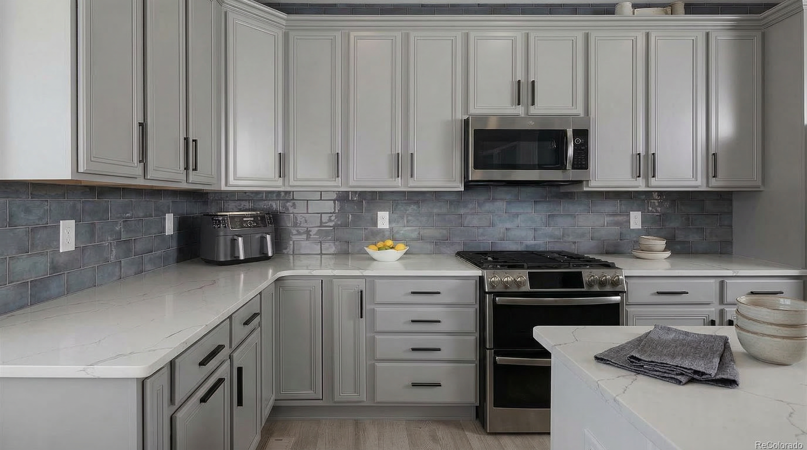 This sleek and modern kitchen features gray cabinetry paired with elegant white quartz countertops and a distinctive blue subway tile backsplash. The space is equipped with stainless steel appliances, including a built-in microwave and a gas range, creating a highly functional and aesthetic culinary workspace.