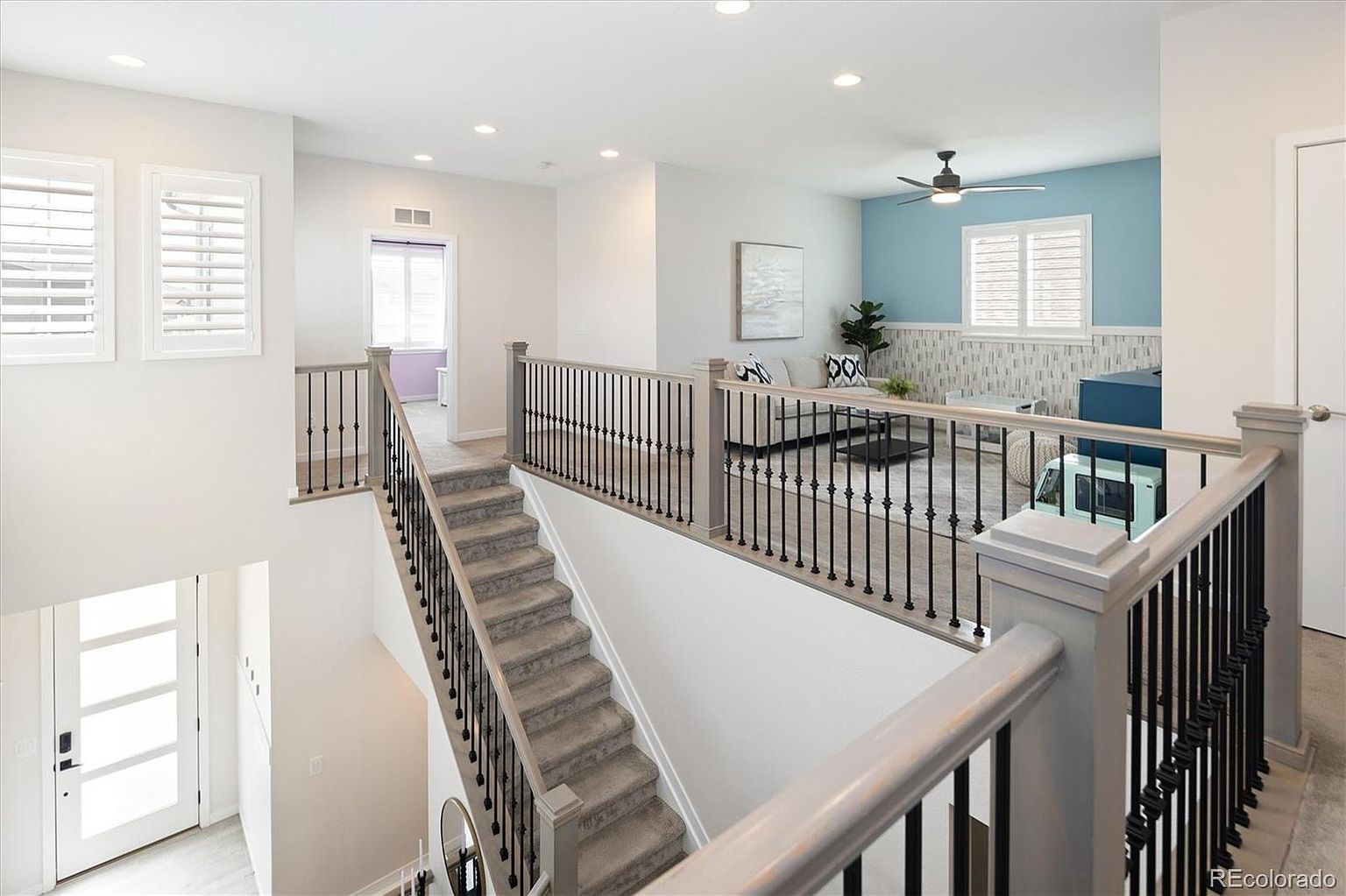 This perspective captures a bright, open-concept landing area featuring a staircase with carpeted steps and elegant black metal balusters. The upper level overlooks a cozy loft space decorated with a blue accent wall, a comfortable sofa, and a playful toy car, creating a welcoming and family-friendly atmosphere. The clean lines, neutral color palette, and recessed lighting contribute to a modern and airy aesthetic throughout the home.