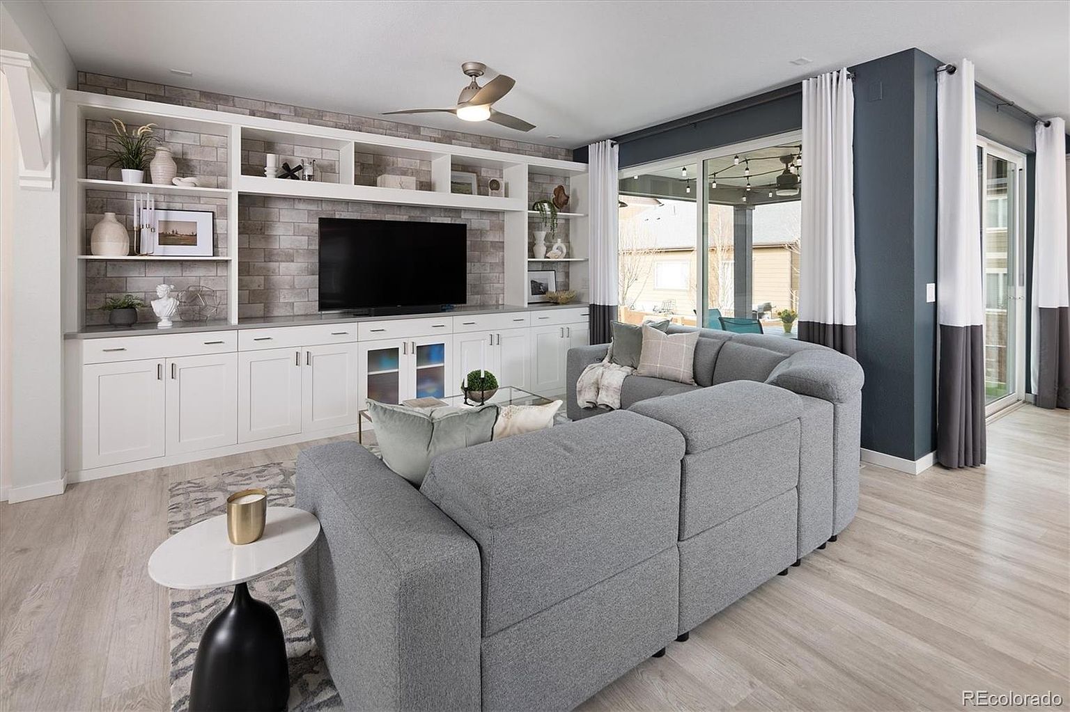 This modern living room features a large, comfortable grey sectional sofa positioned to face a custom built-in entertainment center with a brick-textured backdrop. The space is bright and airy, highlighted by light wood-look flooring, a ceiling fan, and large sliding glass doors that provide a seamless transition to the outdoor patio. The overall aesthetic is contemporary and inviting, perfect for relaxation and entertaining.