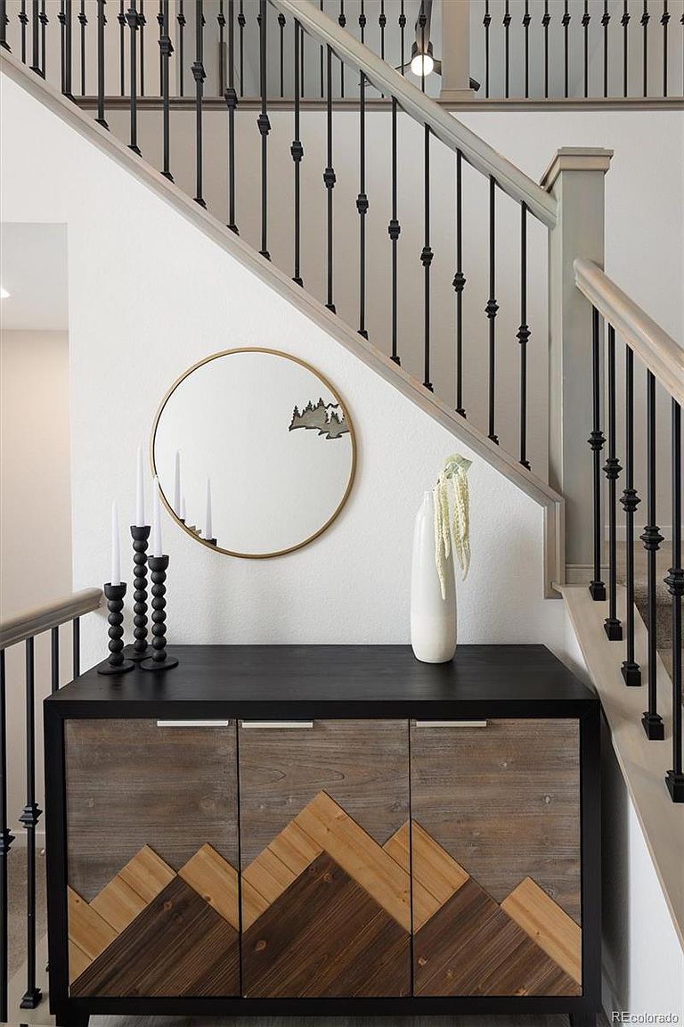 This image captures a stylish landing area at the base of a staircase, featuring a modern console table with a geometric mountain-inspired wood inlay design. Above the table, a circular gold-rimmed mirror reflects the staircase, while a tall white vase and minimalist black candle holders add a sophisticated touch. The scene is framed by elegant black metal railings, creating a clean, contemporary aesthetic that serves as a welcoming focal point.