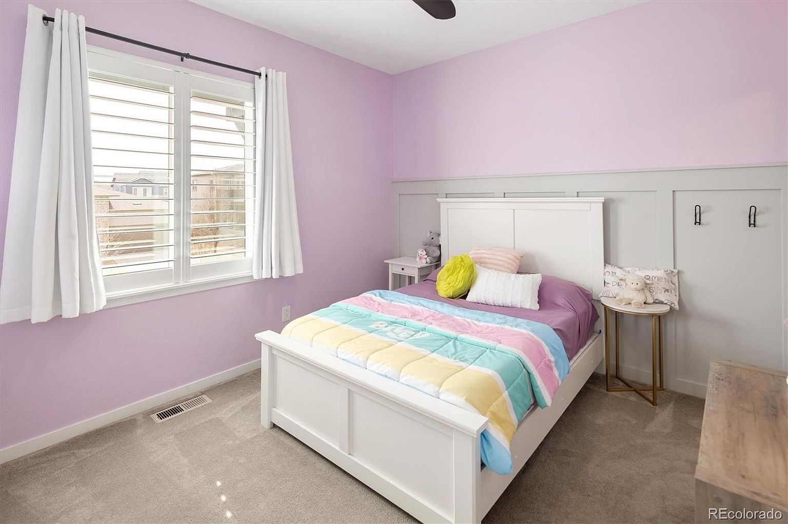 This cozy bedroom features soft lavender walls paired with a white paneled headboard and matching bed frame. A window with white plantation shutters allows natural light to brighten the space, which is furnished with a colorful patterned quilt and a small side table. The room offers a clean, inviting atmosphere suitable for a child's room or guest space.