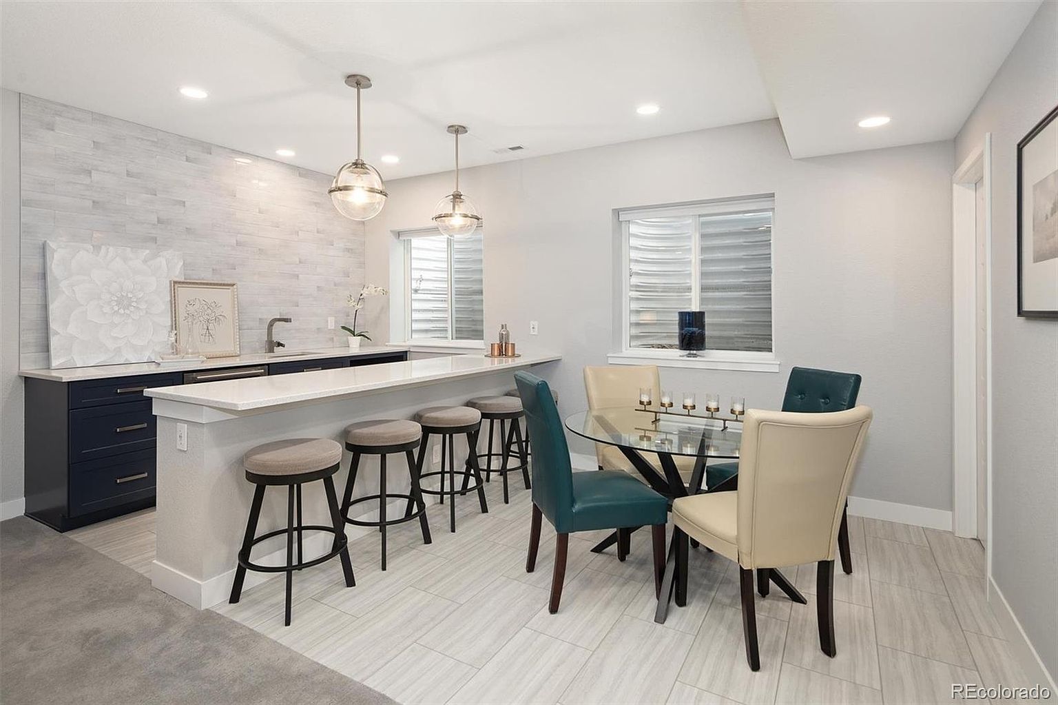 This modern basement space features a sleek wet bar with dark blue cabinetry, a white quartz countertop, and two glass pendant lights. Adjacent to the bar is a cozy dining area with a glass-topped table and a mix of cream and teal upholstered chairs, all set against light wood-look tile flooring. The room is bright and inviting, showcasing a contemporary design perfect for entertaining.