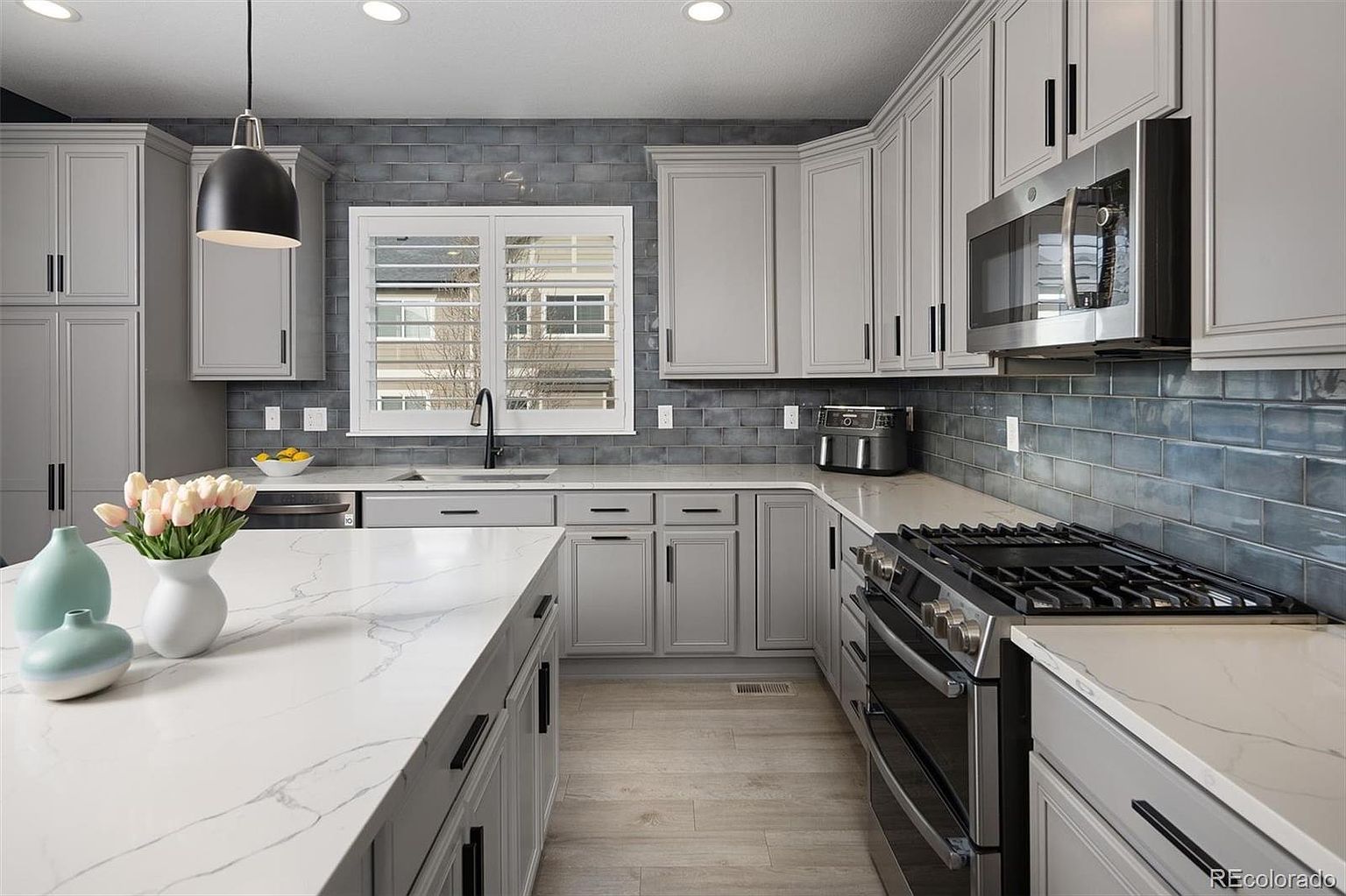 This modern kitchen features sleek light gray cabinetry paired with a striking blue-gray subway tile backsplash and white quartz countertops with subtle veining. A large central island serves as a focal point, complemented by a stainless steel gas range, microwave, and light wood-look flooring. The space is bright and inviting, illuminated by recessed lighting and a stylish pendant light, creating a clean and contemporary aesthetic.