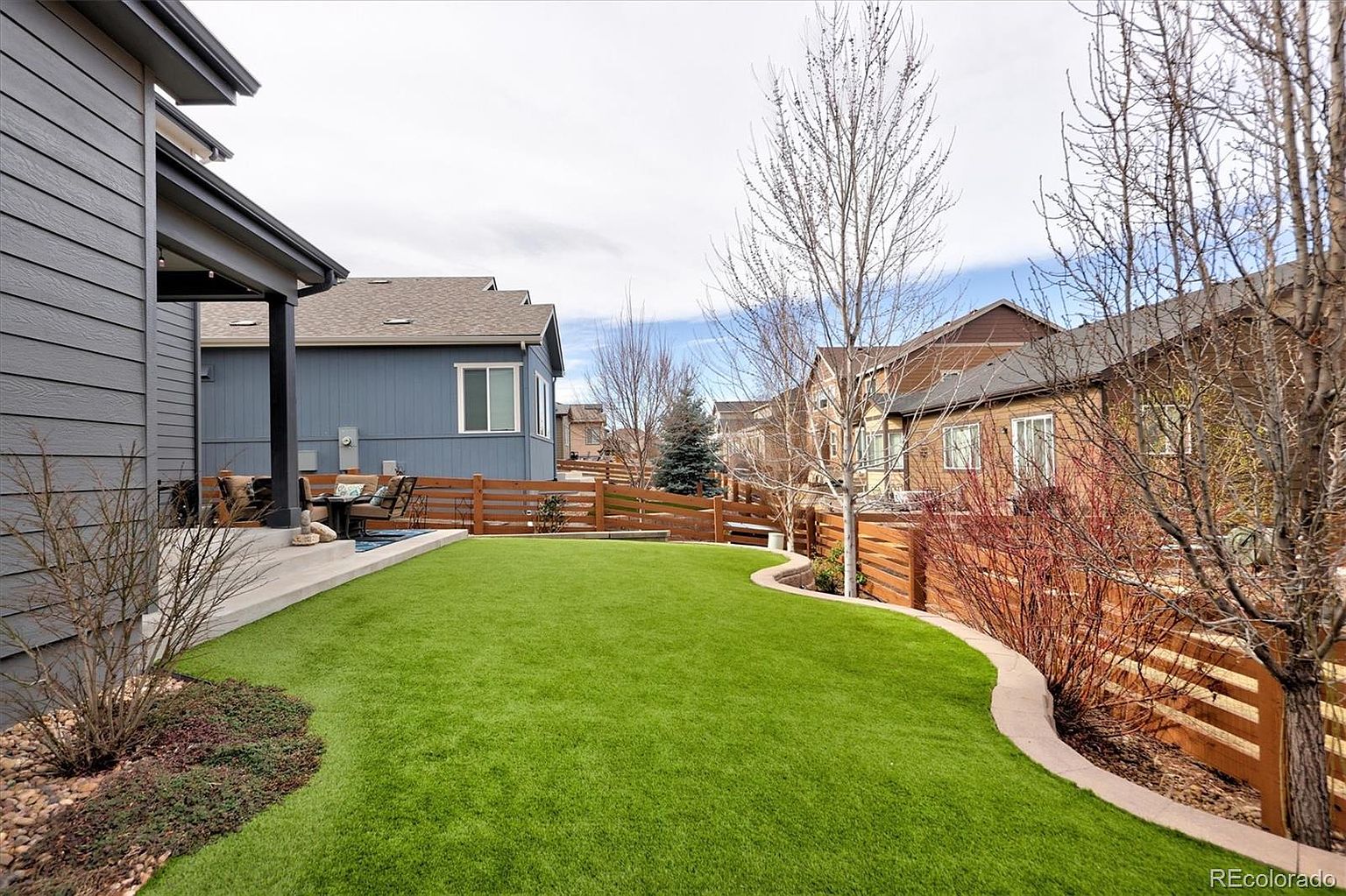 This backyard features a well-manicured artificial turf lawn with a curved stone border, creating a clean and low-maintenance outdoor space. A covered concrete patio area provides a cozy spot for outdoor seating, while a horizontal wood fence offers privacy and a modern aesthetic. The scene is framed by mature trees and neighboring homes, presenting a peaceful and inviting suburban atmosphere.