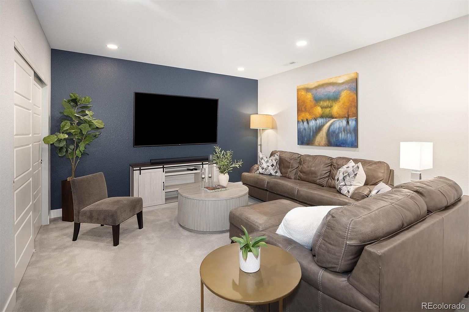 This cozy living room features a striking navy blue accent wall behind a large flat-screen television and a light-toned media console. The space is furnished with a comfortable brown leather sectional sofa, a matching armchair, and a circular coffee table, all set against neutral carpeting. A vibrant landscape painting adds a pop of color to the light-colored walls, creating a warm and inviting atmosphere perfect for relaxation.