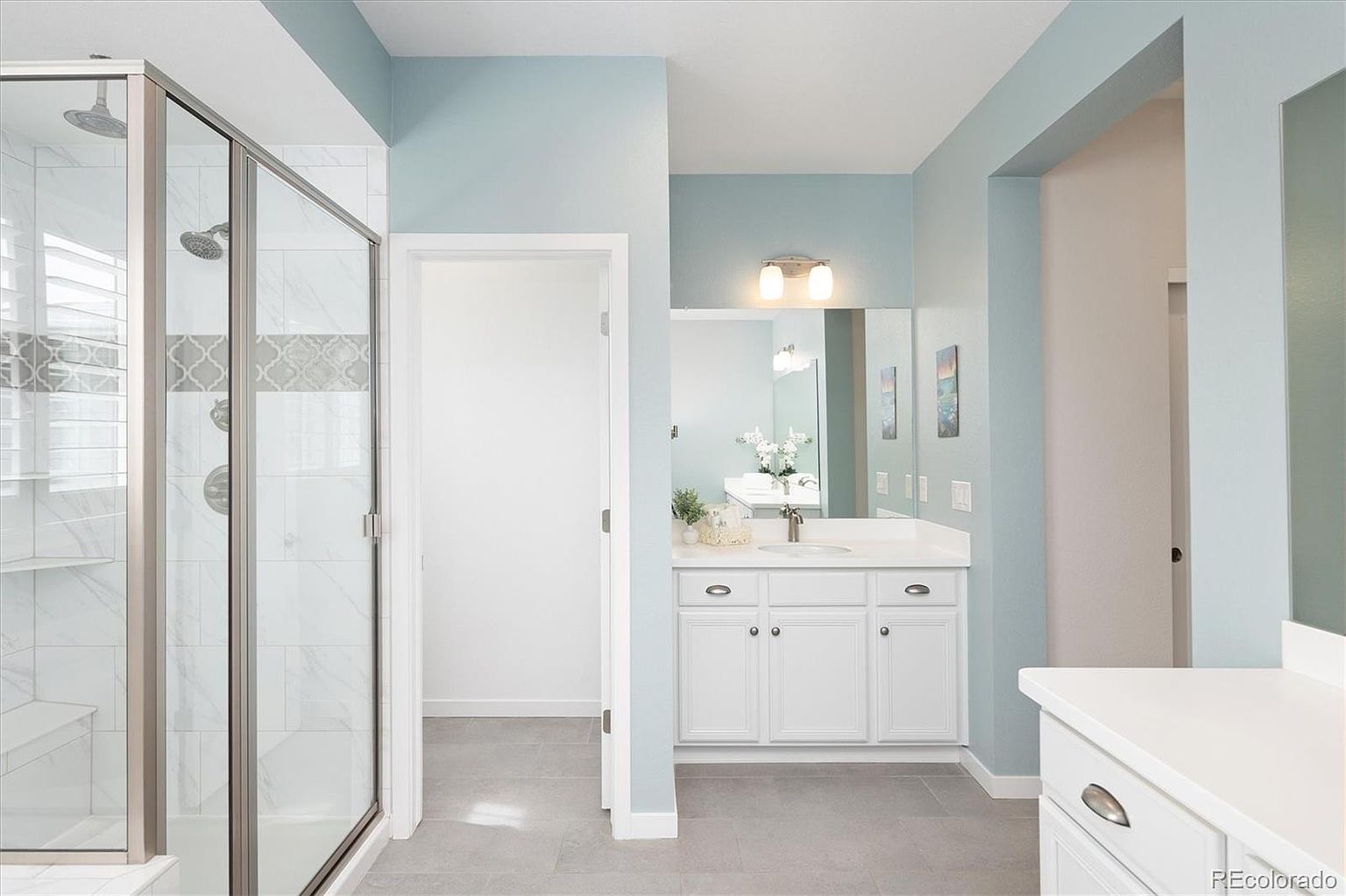 This bright and airy primary bathroom features a clean, contemporary aesthetic with light blue walls and crisp white cabinetry. The space includes a glass-enclosed walk-in shower with marble-style tile accents, a single vanity with a white countertop, and a large mirror that enhances the room's sense of depth. The neutral tile flooring and minimalist design create a serene and functional atmosphere.