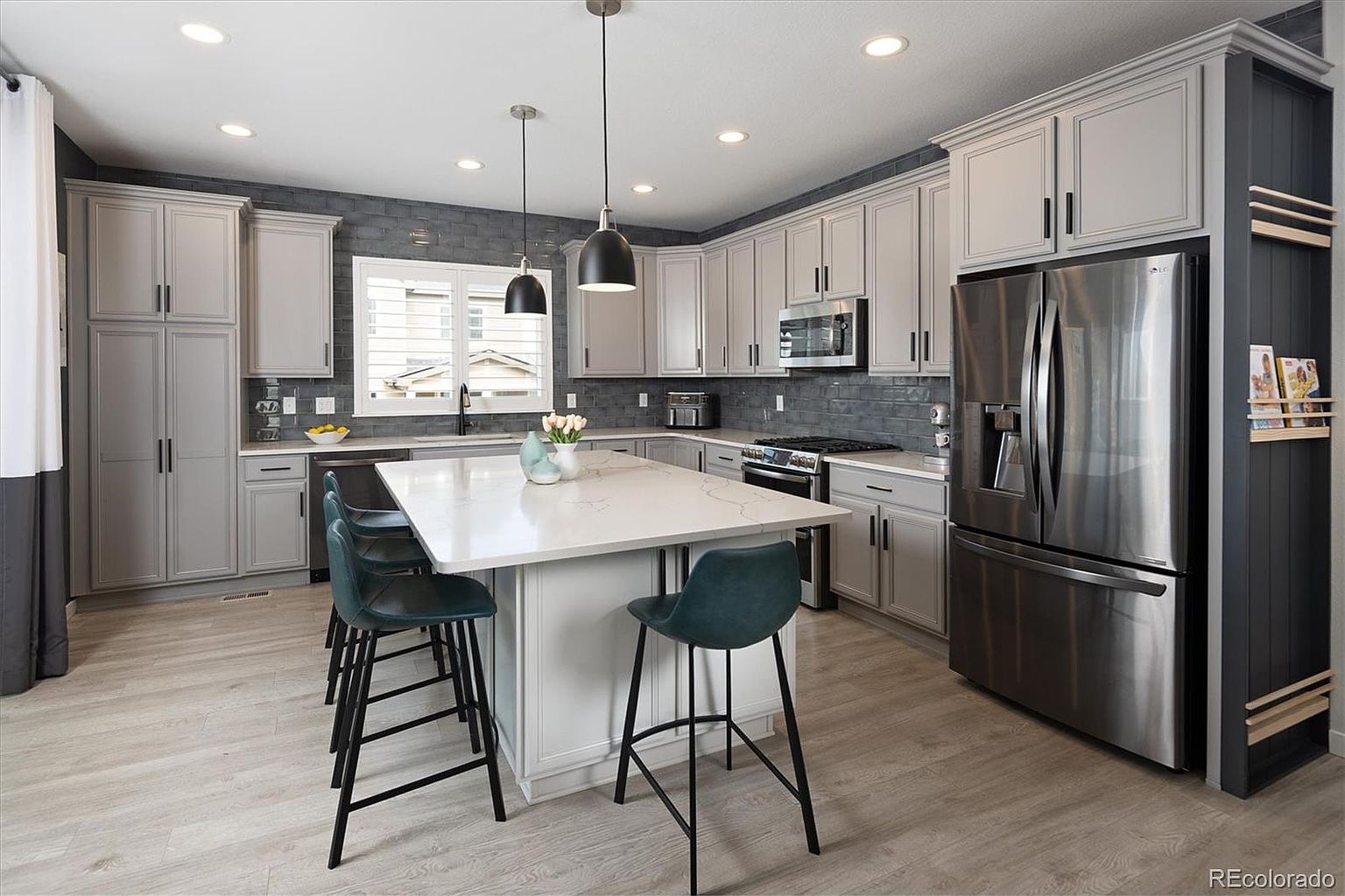 This modern kitchen features light gray cabinetry paired with a dark gray subway tile backsplash and a large central island topped with white quartz. The space is equipped with stainless steel appliances, including a large refrigerator and a gas range, and is illuminated by stylish pendant lighting. The overall aesthetic is clean, contemporary, and functional, with a warm wood-look floor that ties the room together.