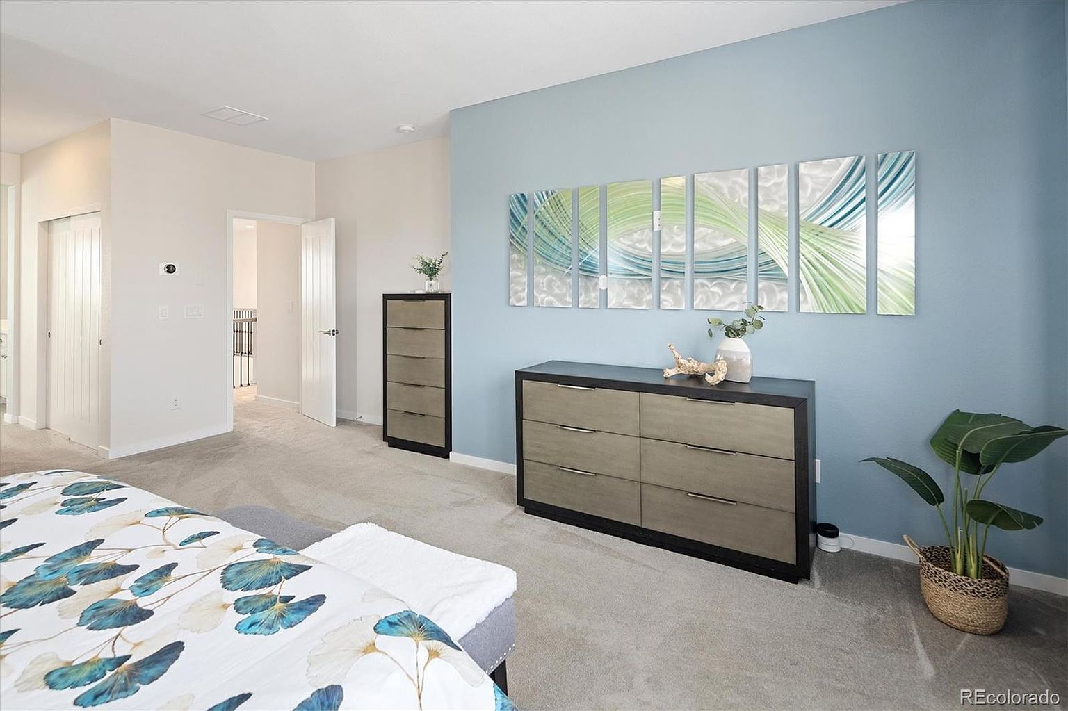This primary bedroom features a calming light blue accent wall adorned with a multi-panel abstract art piece, creating a serene focal point. The room is furnished with a modern, dark-toned dresser and a matching tall chest, complemented by a potted plant and a patterned bedspread. The perspective is from the foot of the bed, showcasing a clean, well-lit space with neutral carpeting and a doorway leading to an adjacent hallway.