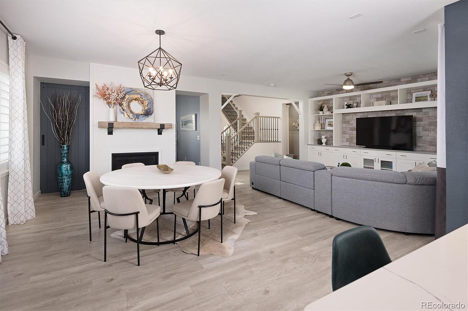 This open-concept living and dining area features a bright, modern aesthetic with light wood-look flooring and a neutral color palette. The space includes a round dining table set against a fireplace with a decorative mantel, while the adjacent living area showcases a large sectional sofa and a custom built-in media wall with shelving. The perspective captures the seamless flow between the dining and living zones, highlighting the home's spacious and inviting atmosphere.