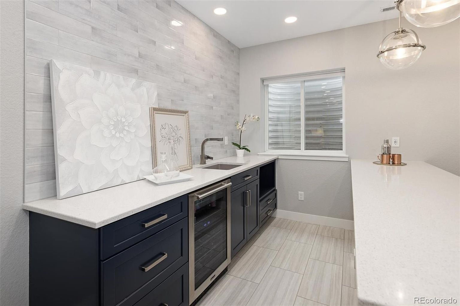 This elegant wet bar area features dark navy cabinetry contrasted with crisp white quartz countertops and a light-toned, linear-tiled backsplash. The space is equipped with a stainless steel wine refrigerator, a sleek undermount sink, and modern pendant lighting, creating a sophisticated and functional entertaining zone. The perspective is a medium-wide shot that highlights the clean lines and contemporary aesthetic of this well-appointed home feature.