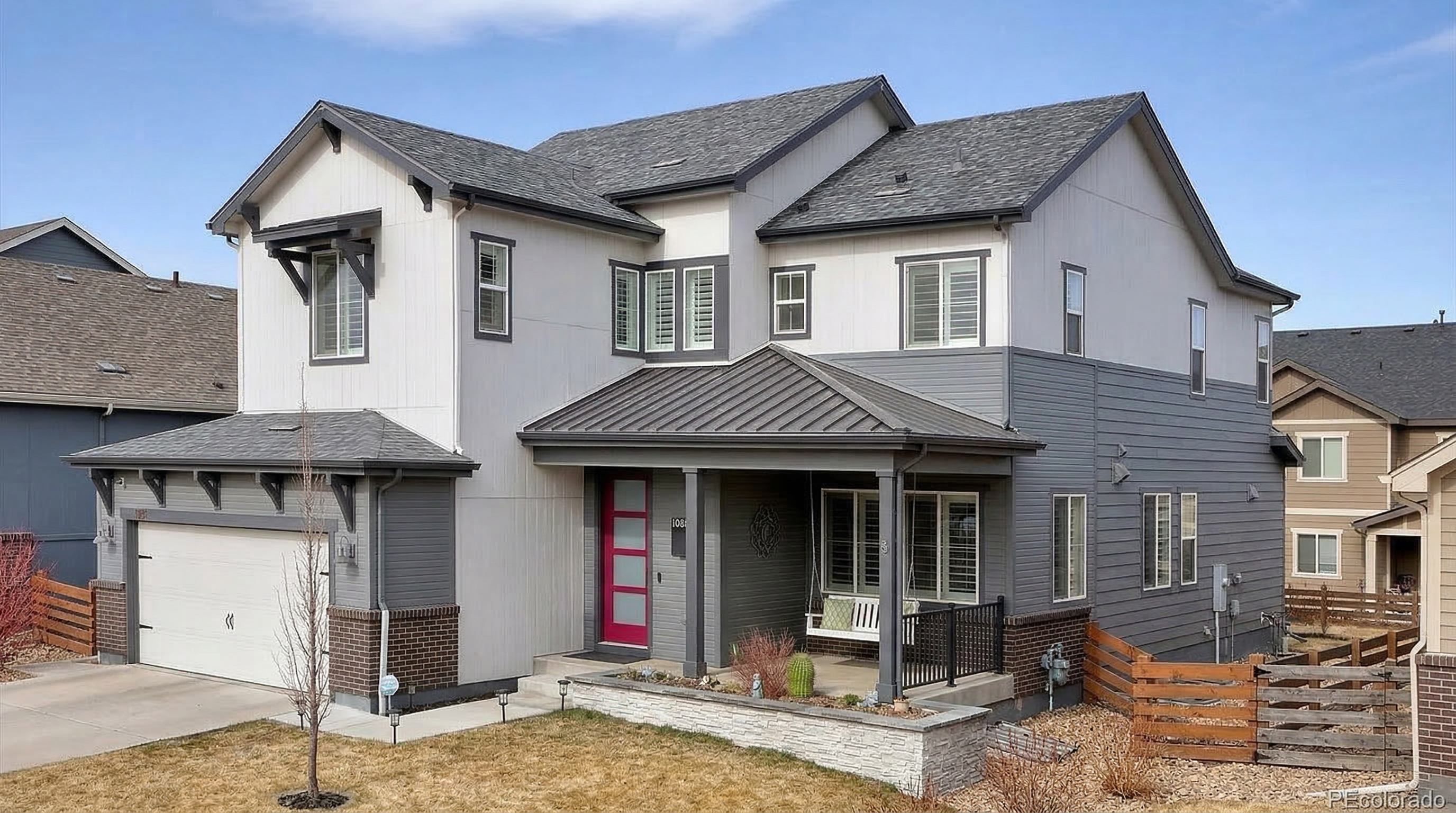 This modern two-story suburban home features a sophisticated blend of grey horizontal siding, white vertical siding, and dark brick accents. The exterior is highlighted by a distinctive, vibrant pink front door, a covered porch with a hanging swing, and a metal-roofed entryway, all contributing to an appealing and contemporary curb appeal.