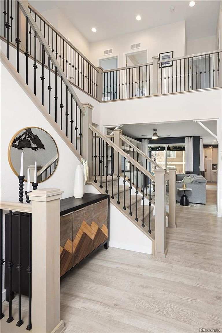 This grand entryway features a striking staircase with black iron balusters and light wood handrails, creating a modern and airy aesthetic. A unique geometric-patterned console table sits beneath a circular mirror, while the open floor plan provides a clear view into the adjacent living area. The light-toned wood flooring and high ceilings contribute to a bright, welcoming, and sophisticated atmosphere.