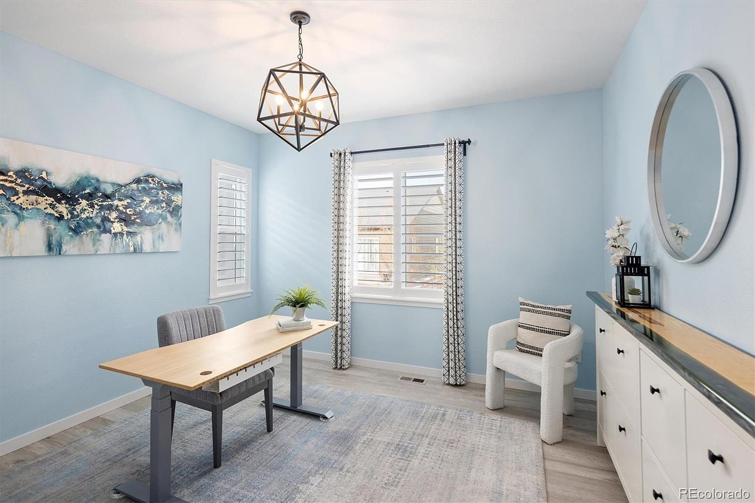 This bright and airy home office features light blue walls, a modern standing desk, and a cozy white accent chair, creating a productive yet inviting atmosphere. A large, geometric pendant light serves as a focal point above the desk, while a circular mirror and abstract wall art add sophisticated decorative touches. The room is grounded by a textured area rug and benefits from natural light streaming through plantation-shuttered windows.