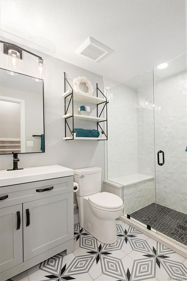 This modern bathroom features a sleek gray vanity with a white countertop, a contemporary black faucet, and a matching framed mirror. The space includes a walk-in shower with a glass door, textured white wall tiles, and a striking geometric patterned floor. A three-tier open shelving unit provides stylish storage, creating a clean, high-end aesthetic.