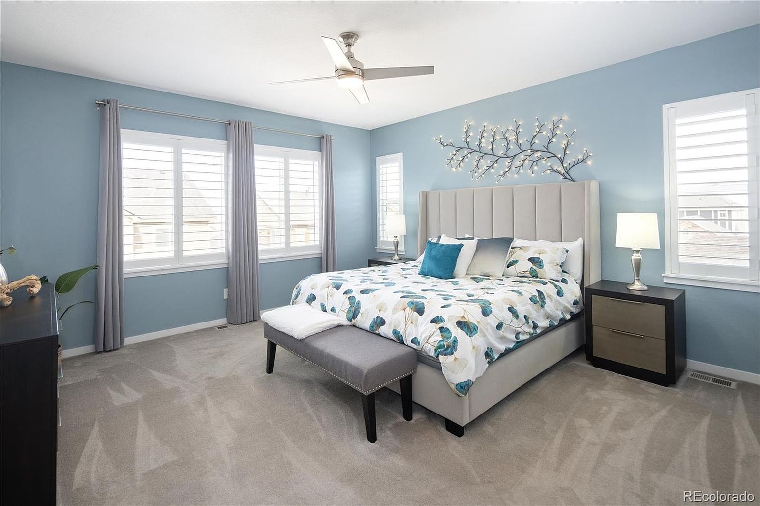 This spacious primary bedroom features soft blue walls, a plush gray carpet, and a large upholstered headboard as the focal point. The room is well-lit by natural light streaming through plantation-shuttered windows and a decorative wall-mounted light fixture above the bed. A matching gray bench sits at the foot of the bed, creating a serene and inviting atmosphere.