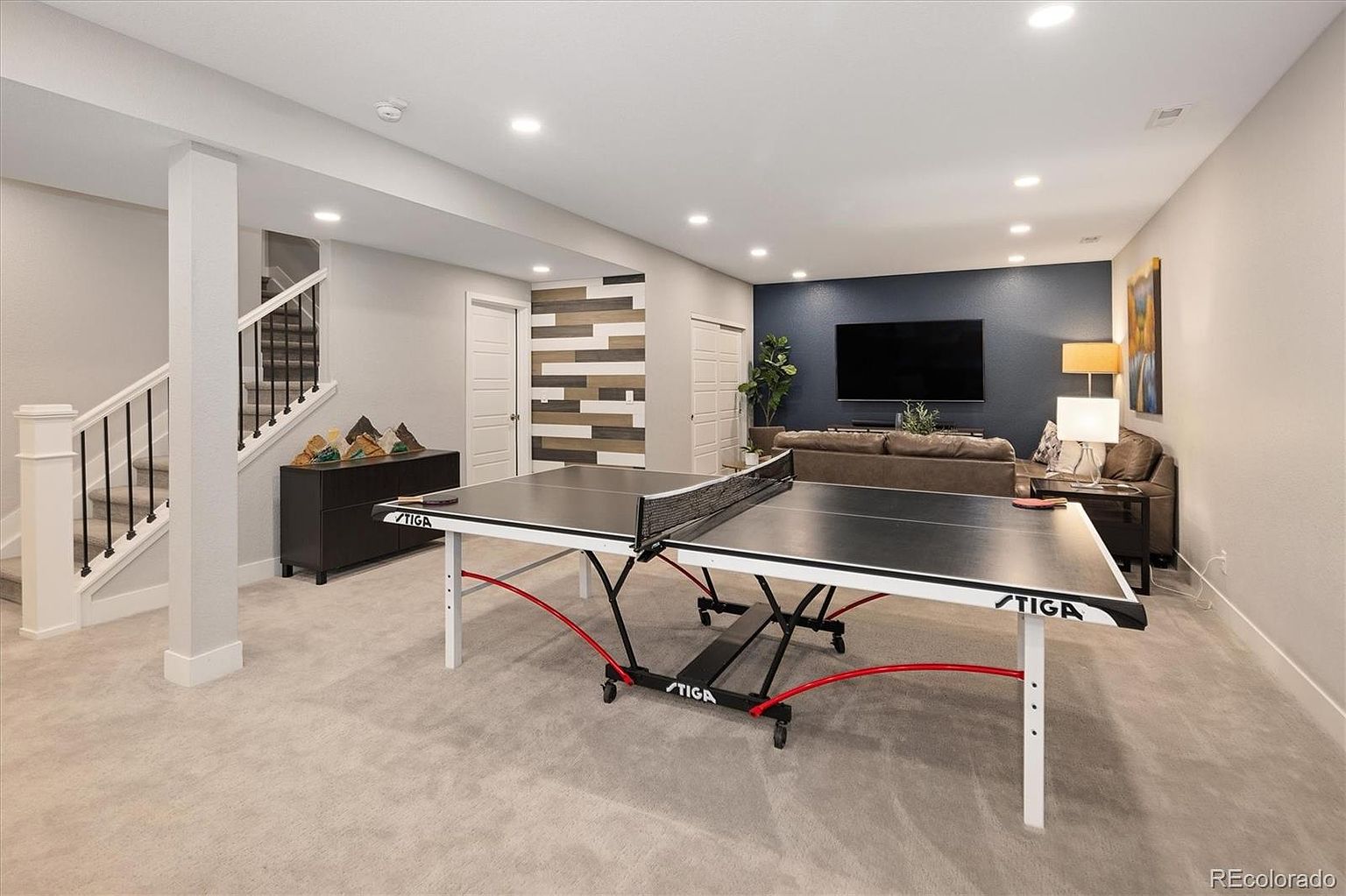 This spacious, finished basement features a neutral color palette with plush carpeting and recessed lighting, creating a comfortable and inviting atmosphere. The room is anchored by a large STIGA ping-pong table in the foreground, while the background showcases a cozy living area with a dark accent wall and a large television. A staircase with white railings and a decorative wood-paneled wall add architectural interest to this versatile entertainment space.