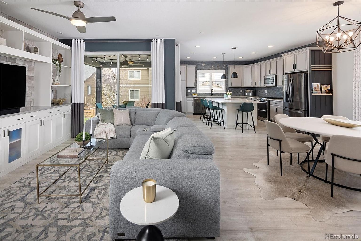 This open-concept living space features a large, plush grey sectional sofa centered around a glass-topped coffee table, set against a backdrop of custom built-in shelving and a television. The room flows seamlessly into a bright, modern kitchen with a central island and a dining area, all unified by light-toned wood flooring. The perspective captures the airy, cohesive design of the home, highlighting the transition between relaxation and culinary spaces.