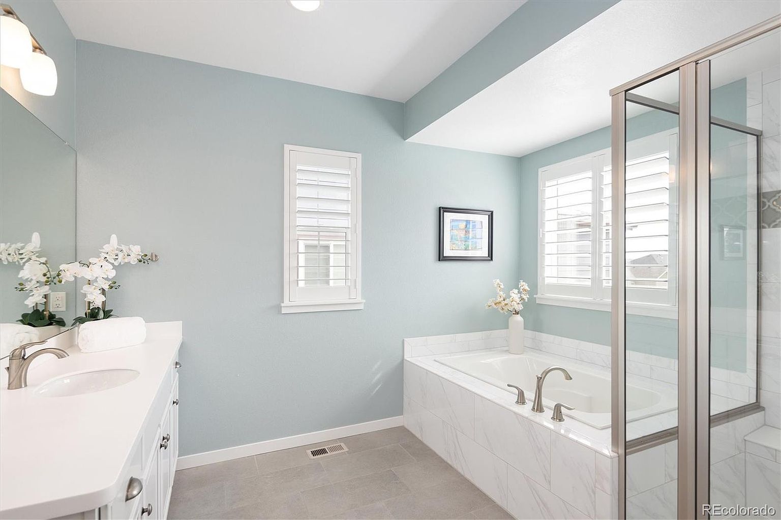 This bright and airy primary bathroom features a clean, modern aesthetic with light blue walls and white marble-tiled accents. The space includes a spacious vanity with a white countertop, a deep soaking tub surrounded by marble, and a glass-enclosed shower. The room is well-lit by natural light from the windows and elegant vanity lighting, creating a serene and spa-like atmosphere.