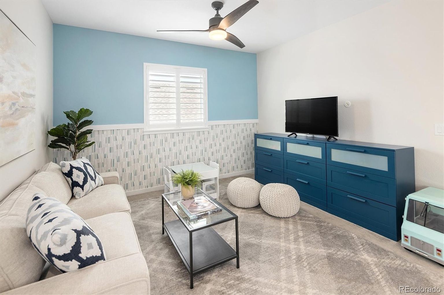 This cozy and playful living space features a light-colored sofa adorned with patterned throw pillows, a glass-topped coffee table, and a vibrant blue dresser serving as a media console. The room is accented by a two-toned wall with patterned wallpaper, a ceiling fan, and a soft gray area rug, creating a welcoming and family-friendly atmosphere. A small child's table and chair set, along with a toy vehicle, suggest a versatile space designed for both relaxation and play.