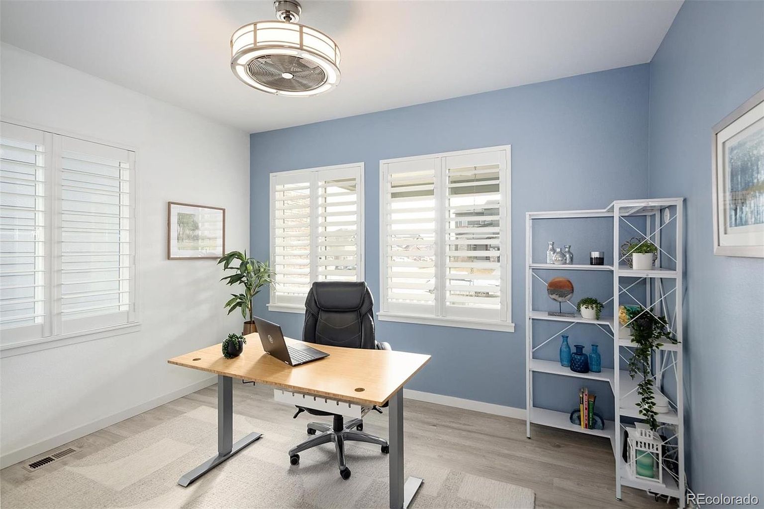 This bright and modern home office features a light wood-topped standing desk, a comfortable black ergonomic chair, and a white open-shelving unit decorated with plants and decor. The room is accented by a soft blue feature wall, white plantation shutters on the windows, and light-toned wood flooring, creating a clean and professional atmosphere. The perspective is a straight-on, eye-level shot that highlights the functional layout and inviting aesthetic of the workspace.