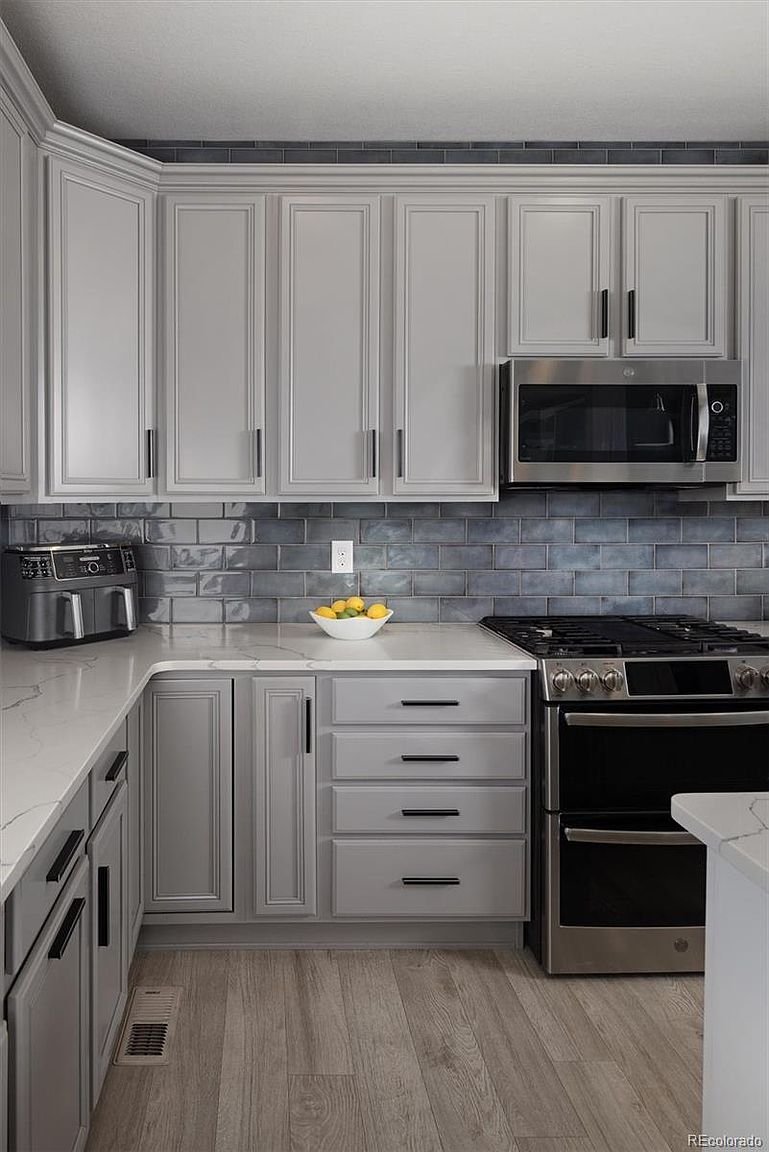 This modern kitchen features sleek light gray cabinetry paired with a striking dark blue subway tile backsplash and white quartz countertops. The space is equipped with stainless steel appliances, including a double oven range and an over-the-range microwave, creating a clean and functional aesthetic. The light wood-look flooring adds warmth to the contemporary design, making it an inviting and well-organized culinary space.