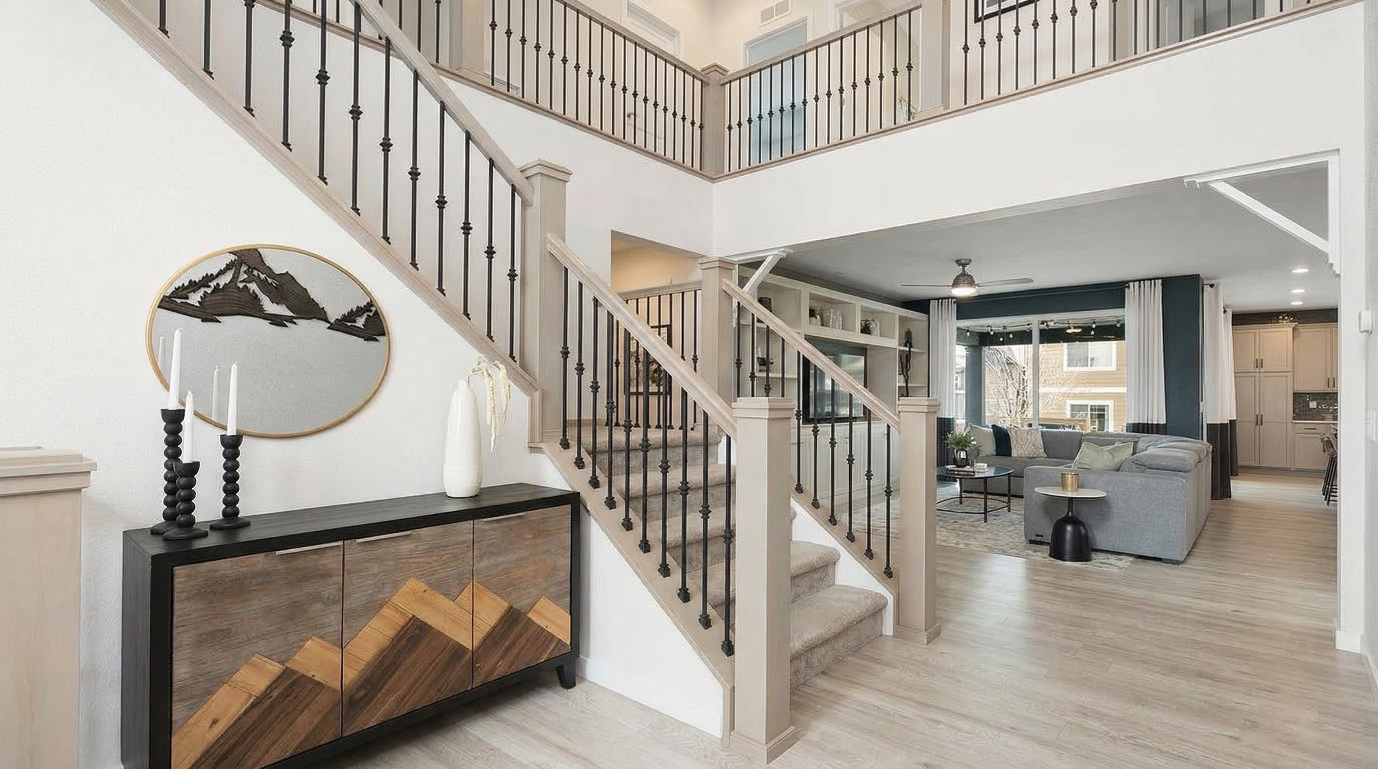 This grand entryway features an elegant staircase with wood railings and black iron spindles, leading to a sprawling open-concept main floor. The space is highlighted by a custom mountain-themed sideboard, a matching circular wall art piece, and light wood-style flooring that flows throughout the home. The design creates a sophisticated, cohesive transition into the living room and kitchen area.