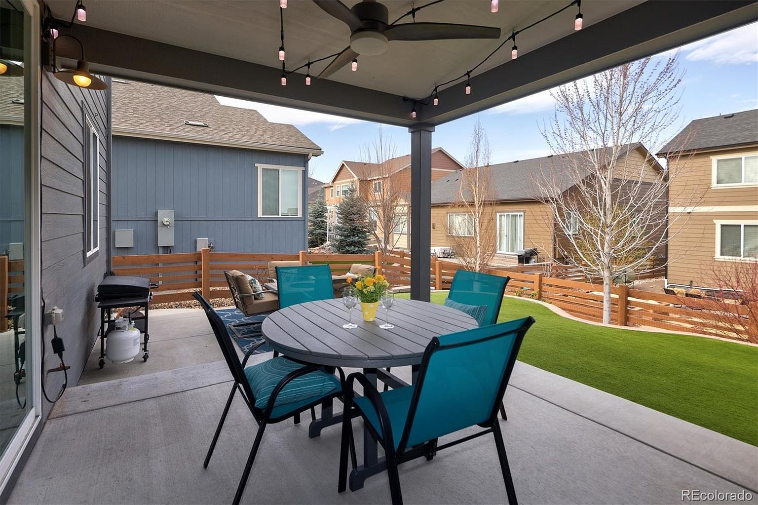 This covered patio offers a comfortable outdoor living space, featuring a round dining table with four teal chairs and a small seating area in the background. The concrete floor extends out to a well-maintained artificial turf lawn, enclosed by a modern horizontal wood fence. The perspective is from under the covered patio, looking out toward the neighboring homes, creating an inviting and private atmosphere for outdoor dining or relaxation.