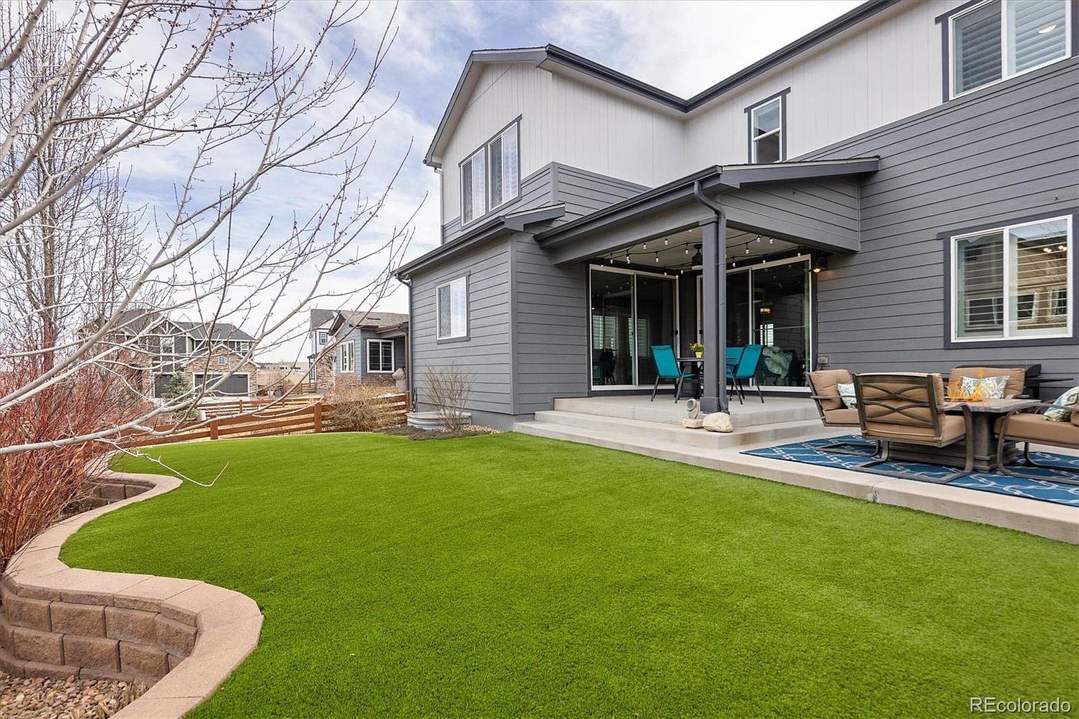 This image showcases a well-maintained backyard featuring a vibrant green artificial turf lawn and a concrete patio area furnished with outdoor seating. The rear of the two-story home is finished in gray horizontal siding with white trim, highlighting a covered patio space illuminated by string lights. The perspective captures the inviting outdoor living space, emphasizing the transition from the manicured lawn to the comfortable patio area.