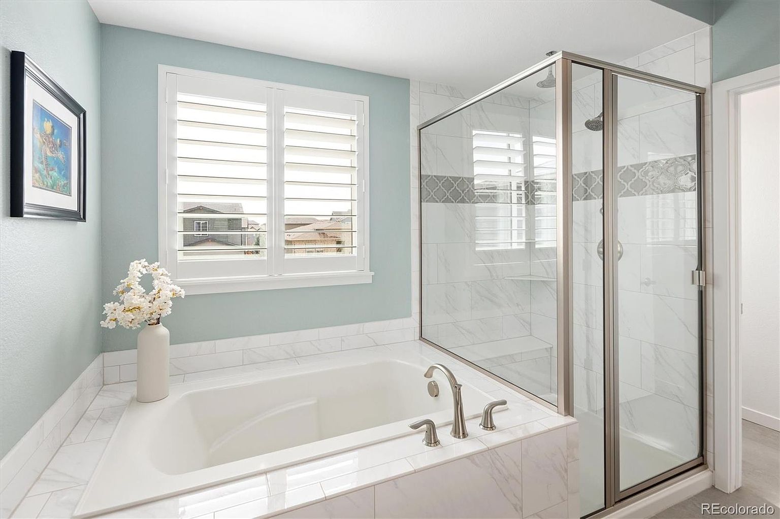 This bright and airy primary bathroom features a deep soaking tub set against a light blue wall with plantation shutters. Adjacent to the tub is a spacious walk-in glass shower with marble-look tile and a decorative accent border. The clean, neutral color palette and modern fixtures create a serene and spa-like atmosphere.
