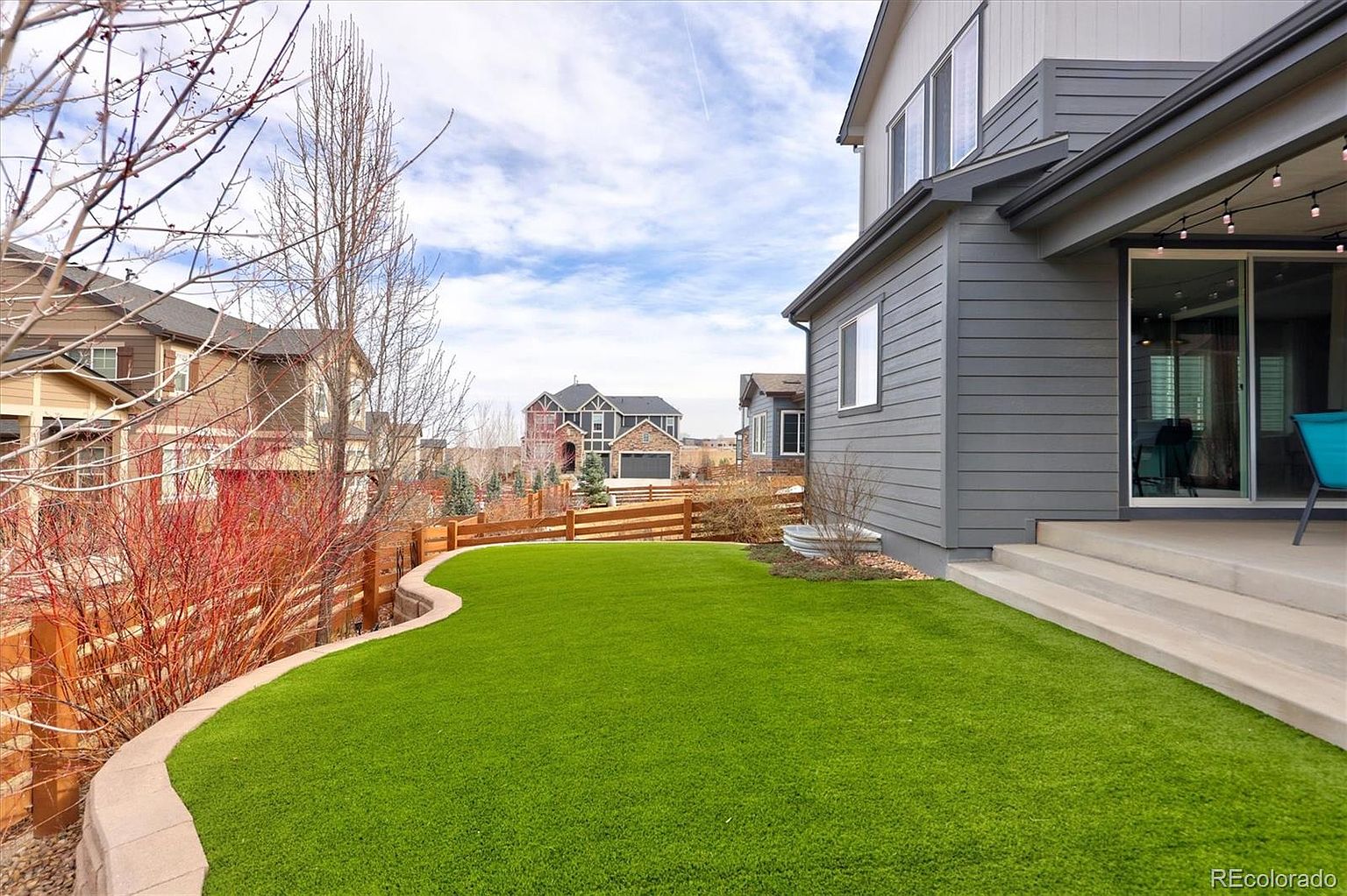 This image showcases a well-maintained backyard featuring a vibrant green artificial turf lawn bordered by a curved stone retaining wall. A modern gray-sided house with a concrete patio and string lights provides a comfortable outdoor living space, while a wooden fence separates the property from neighboring homes. The perspective captures the inviting, low-maintenance landscape under a bright, partly cloudy sky.