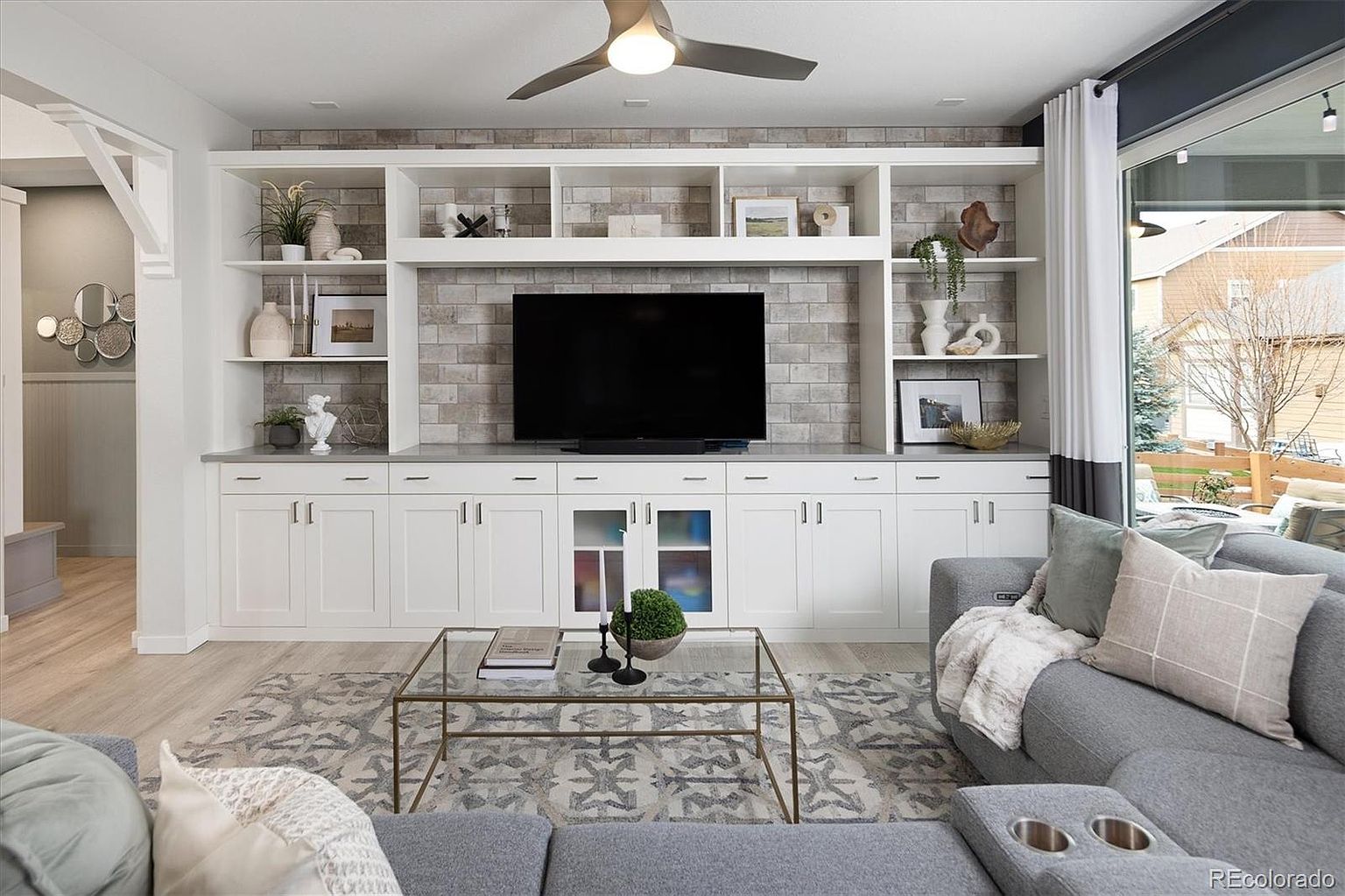 This bright and inviting living room features a prominent custom built-in entertainment center with white cabinetry and a textured brick backsplash. A large, comfortable grey sectional sofa faces the television, complemented by a glass-topped coffee table and a patterned area rug. The space is illuminated by a modern ceiling fan and natural light streaming in from the large window, creating a warm and contemporary atmosphere.